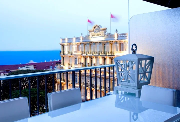 Hotel Hermitage building seen from a balcony with a table, chairs, and a lantern in the foreground, overlooking the ocean.