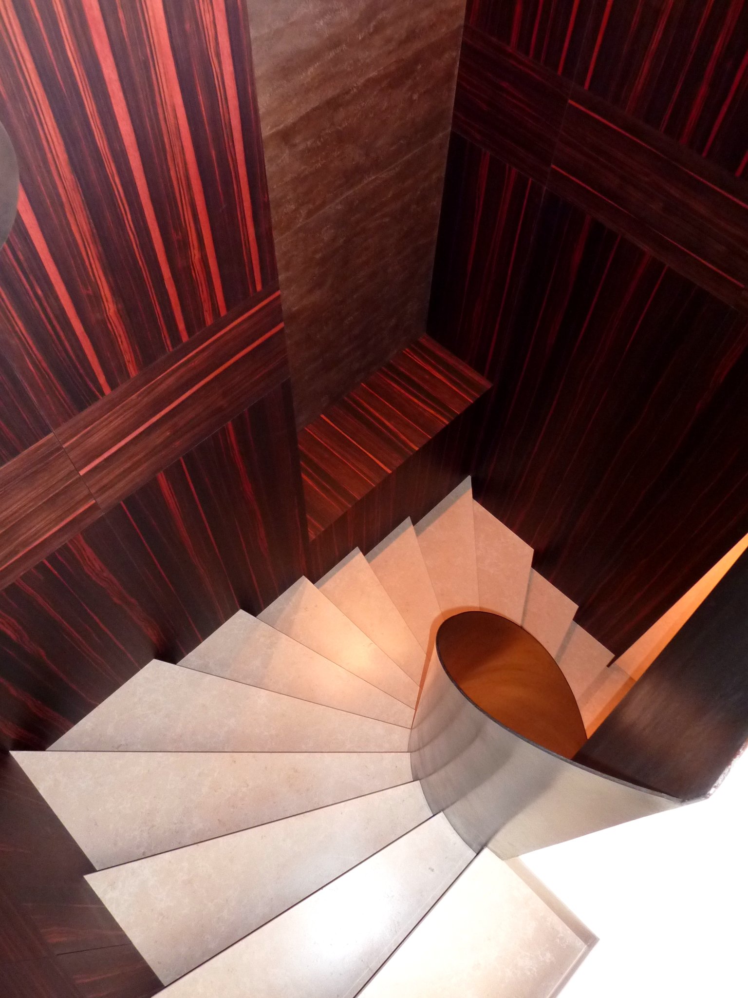 An overhead view of a spiral staircase with beige steps, a wooden handrail, and dark wood paneling on the walls.