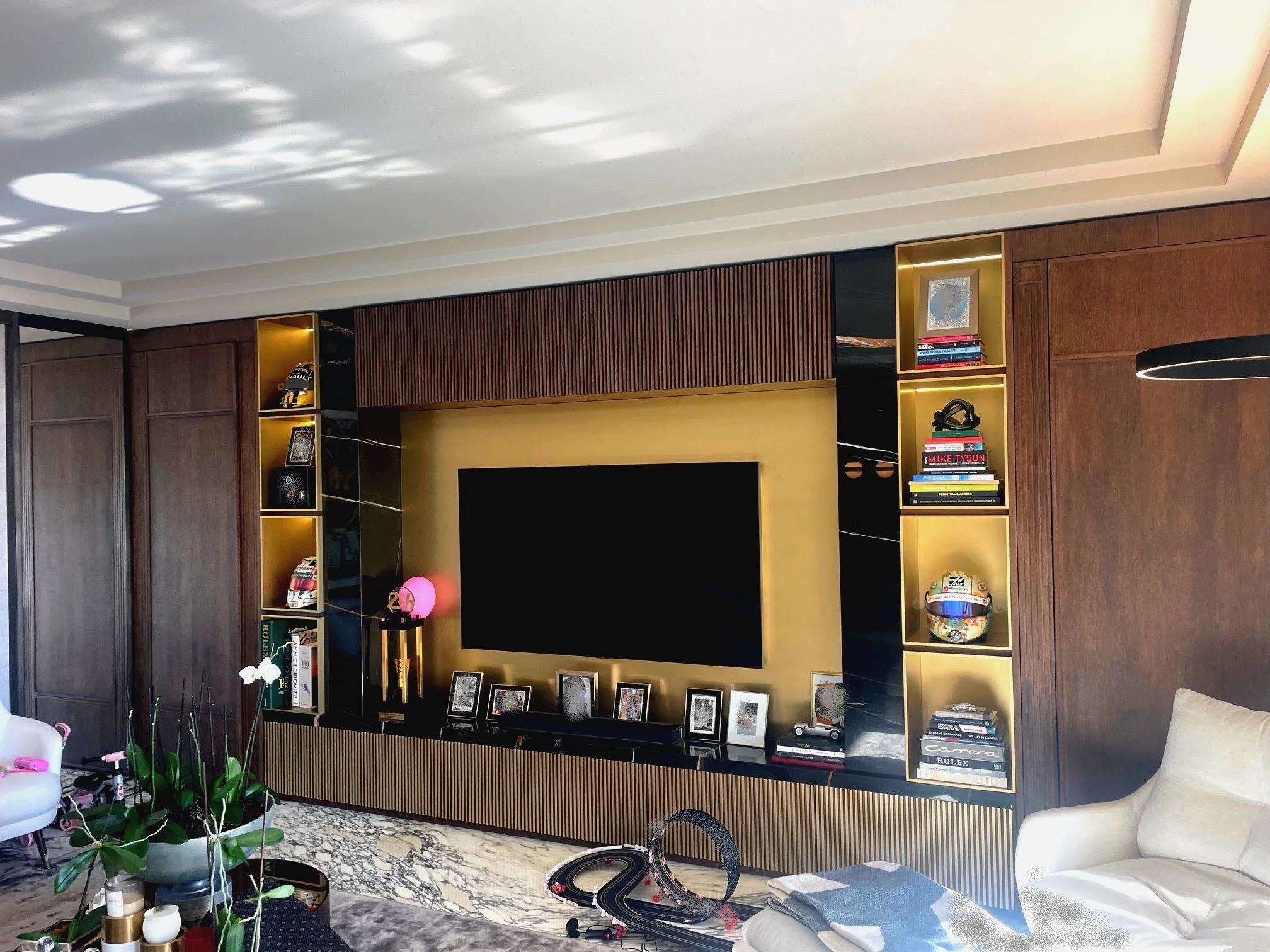 Modern living room featuring a wall-mounted flat screen TV, built-in bookshelf with decorative items, framed photos, books, and a toy car. There is a beige sofa, potted plants, and a black and gold entertainment console on a marble floor.