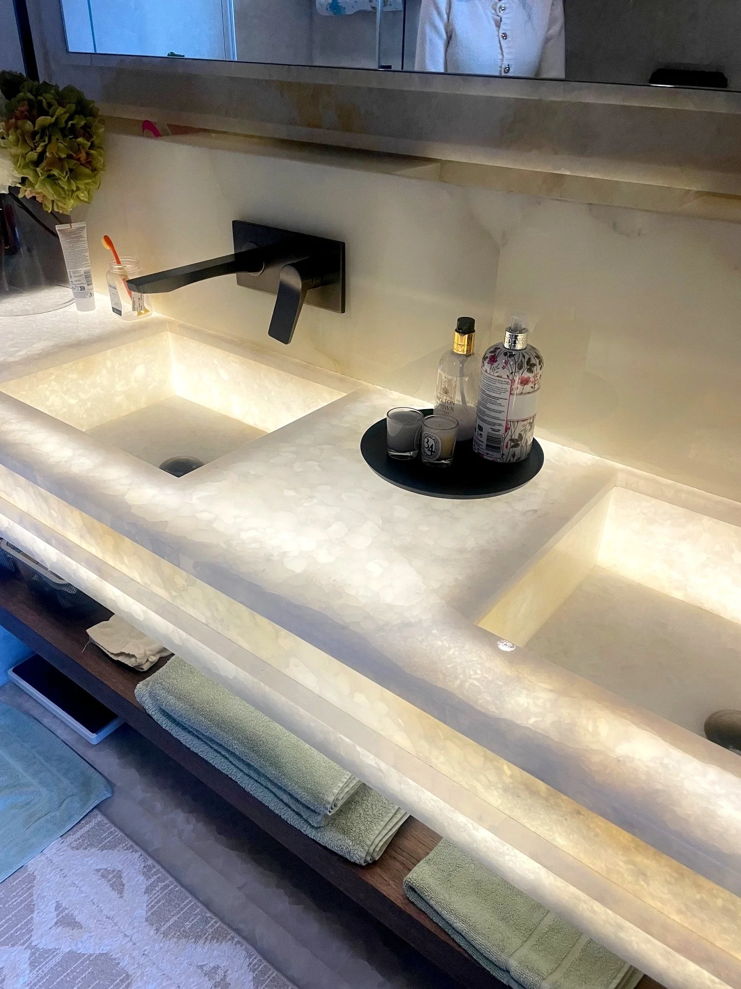 A illuminated bathroom sink with a rectangular basin and a black faucet, set in a marble countertop with some bottles and candles on a tray nearby. There are towels stored on a shelf underneath.