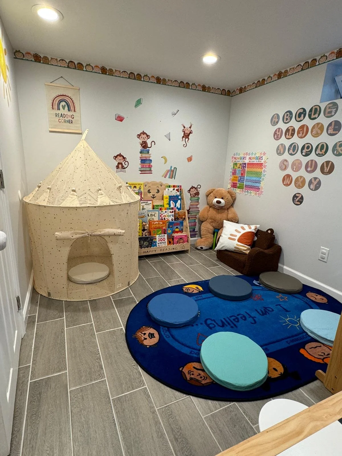 A children's reading and playroom with a cozy tent, a plush teddy bear, a small armchair, colorful cushions on a blue rug, and educational wall decorations including ABCs and numbers.