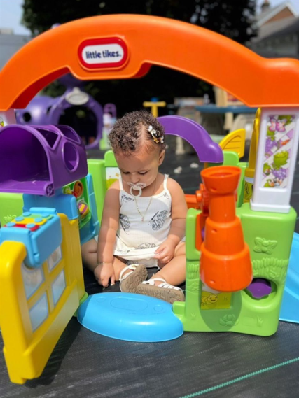 Young child sitting inside a colorful Little Tikes playhouse with purple, orange, green, and blue plastic features, holding a pacifier and sitting on a blue mat outside.