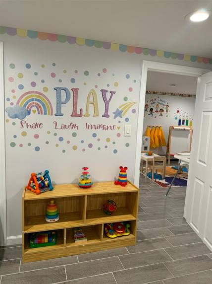 Decorated children's playroom wall with colorful dots, rainbows, stars, and the words 'PLAY' and 'Share Laugh Imagine'; wooden shelves with colorful stacked toys and a red car, and a doorway leading to another play area.