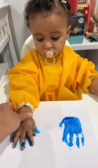 A young child with curly hair wearing a yellow smock and a pacifier in her mouth, sitting at a table. Her hand is being held and pressed onto a piece of white paper with her blue-painted handprint on it.