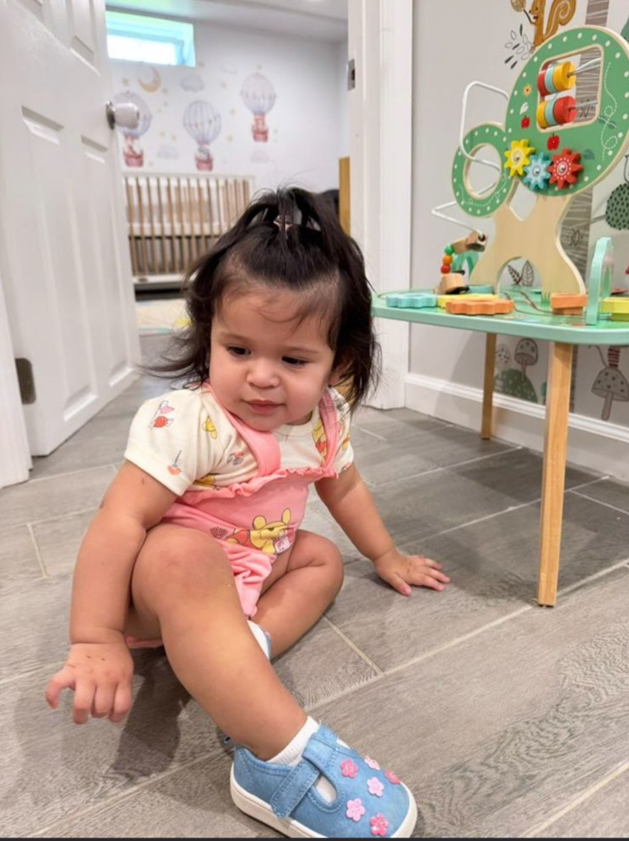 A young girl sitting on a gray tiled floor in a playroom, looking down with a slightly confused expression, wearing a pink dress with a yellow bow design, blue shoes with pink flowers, and white socks. There are children's toys and wall decorations i
