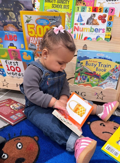 A young girl sitting on a blue carpet with animal designs, looking at a book with an orange and white illustration, surrounded by colorful children's books and educational materials.
