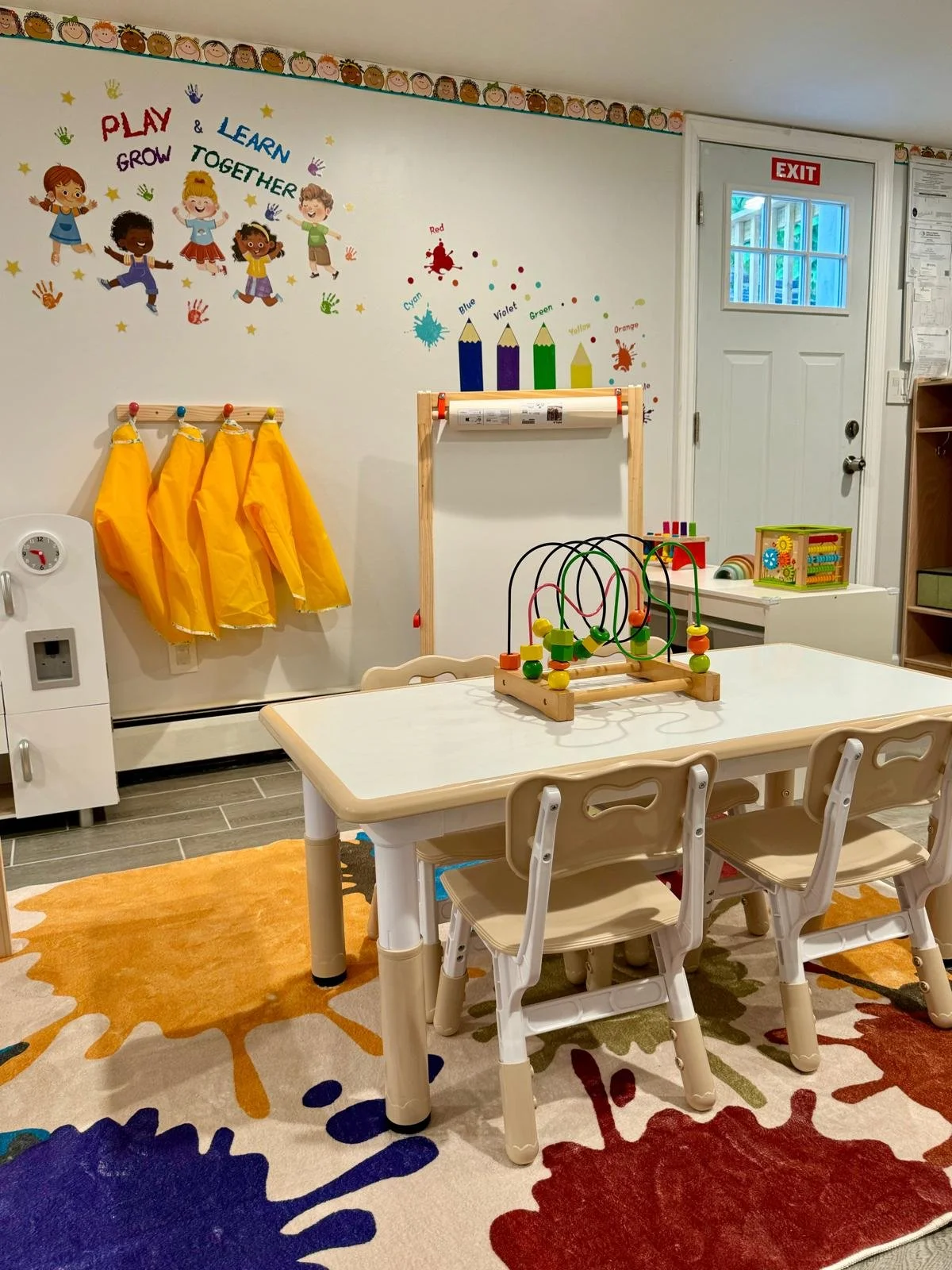 A classroom with a small table and chairs, colorful wall decorations including children illustrations, a calendar, a wall with a chair rail, yellow aprons hanging on hooks, a rainbow-colored rug, and educational toys on shelves.