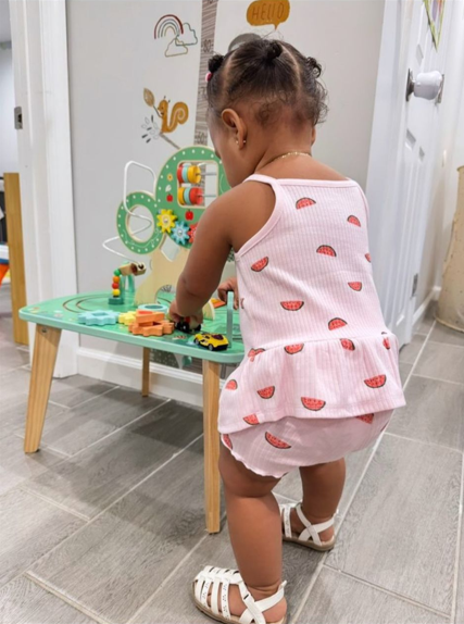A young girl in a white watermelon print romper playing with a toy car and building blocks at a small wooden table in a home.