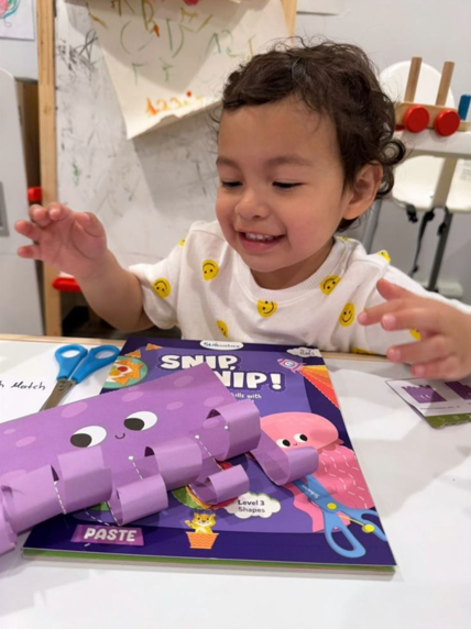 Young child smiling at a craft project on a table, with a purple paper craft shaped like a crab, scissors, and a craft magazine.