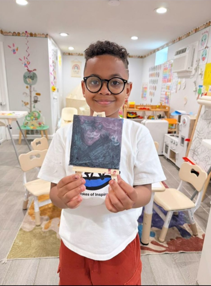 Young boy with glasses holding a small canvas painting in a colorful classroom.
