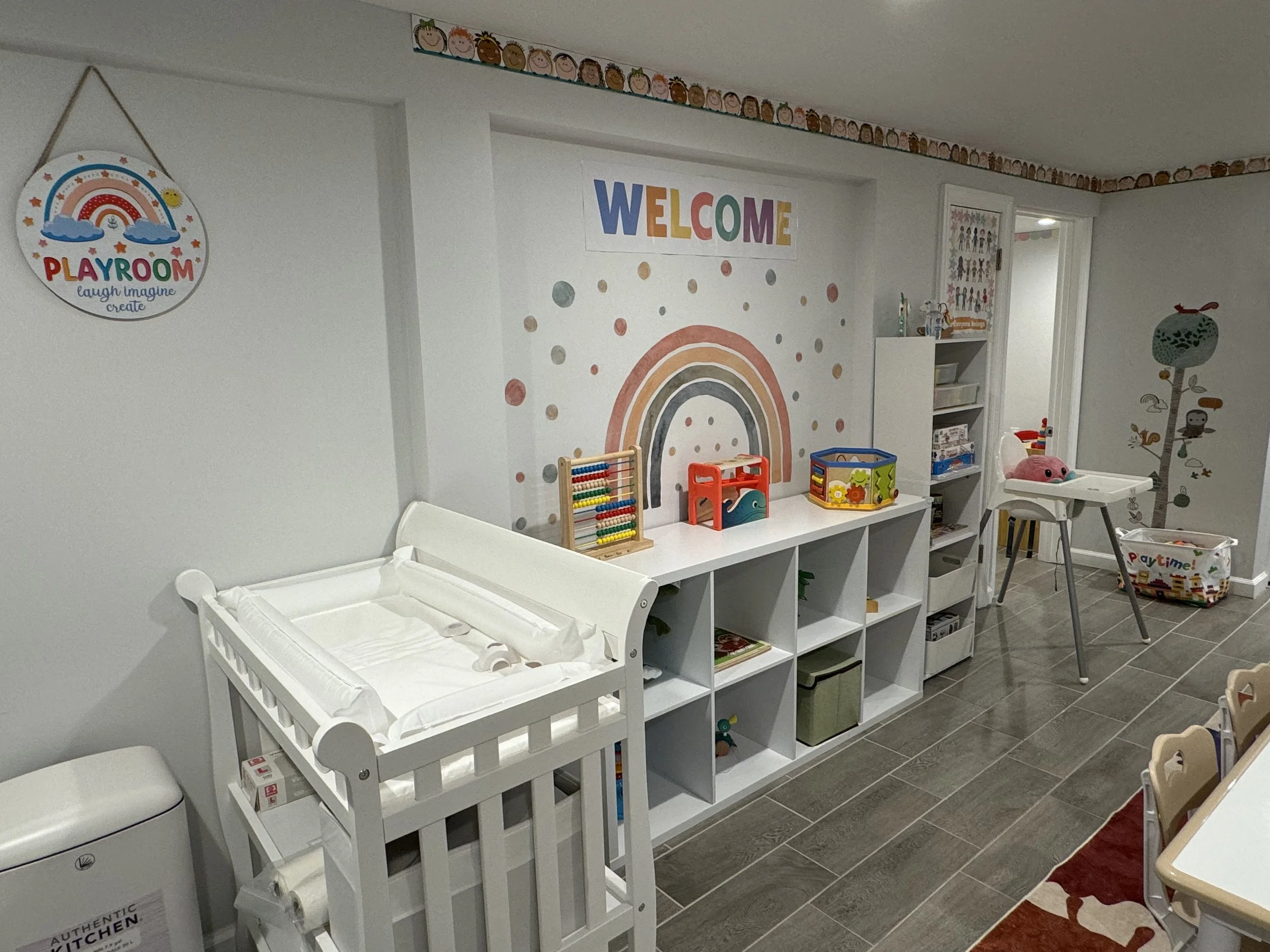 Colorful children's playroom with a white storage cube, a white changing table, wall decorations including a rainbow and a 'PLAYROOM' sign, toys, and a high chair.