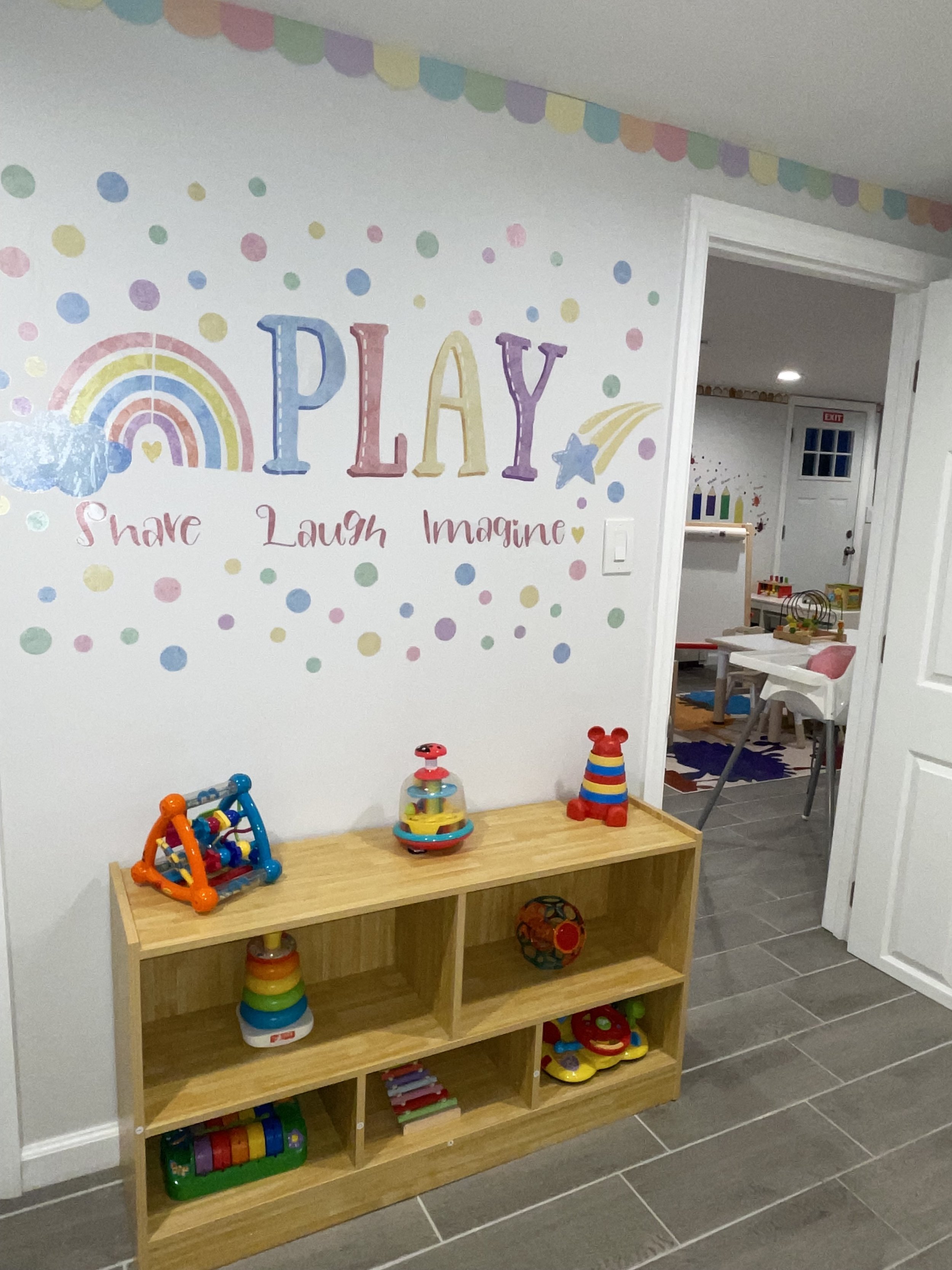 Colorful children's playroom with a painted wall that says 'PLAY' and the words 'Share, Laugh, Imagine' underneath. There is a wooden shelf filled with toys and a doorway leading to another play area with more toys and furniture.