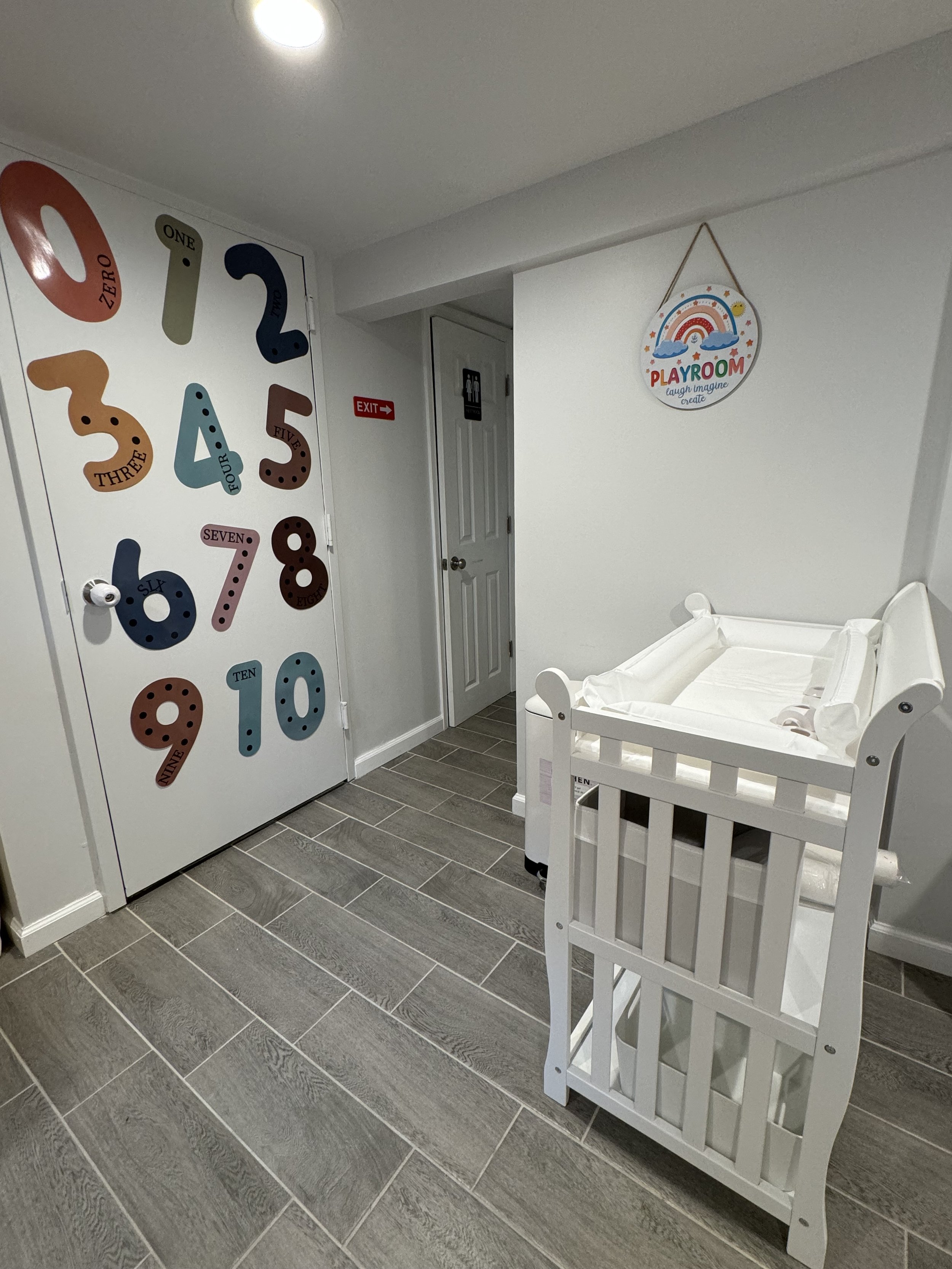 Baby crib in a playroom with a colorful number door and a sign that reads 'Playroom, laugh, imagine, create' hanging on the wall.