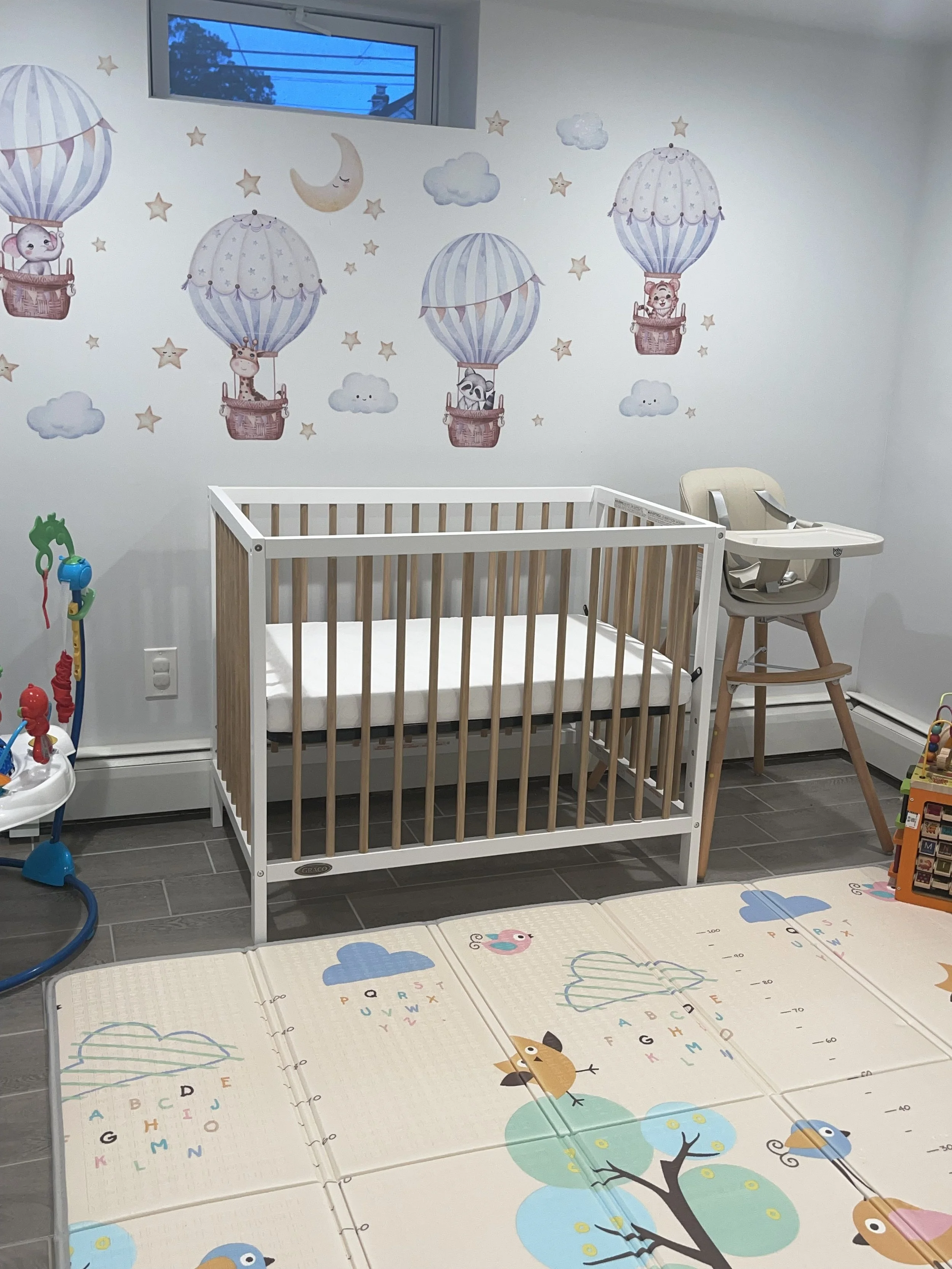 Nursery room with a white and wooden crib, a high chair, and a play mat with colorful birds and clouds, decorated with clouds, stars, moon, and hot air balloon wall decals.