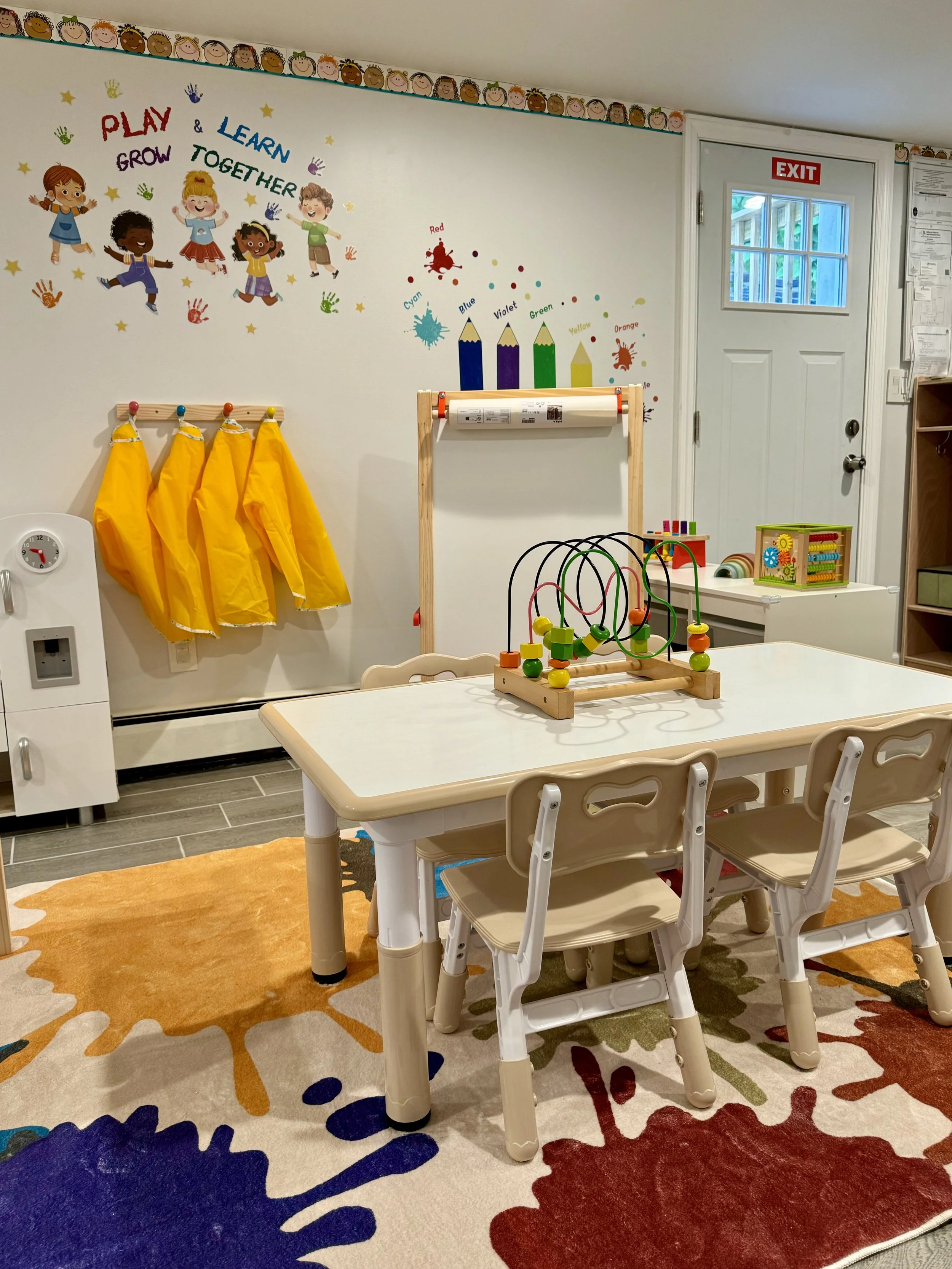 A children's play and learning area with a colorful wall decoration featuring children and text that says "Play, Learn & Grow Together." There are yellow smocks hanging on hooks, a table with toys including a bead maze, a colorful rug with splashes o