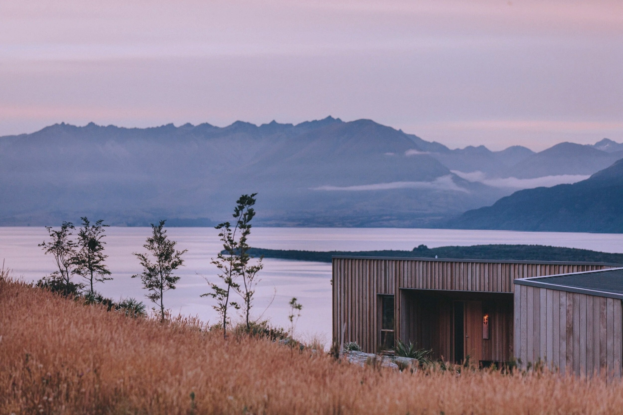 A modern wooden house with large windows on a hillside with tall grasses, overlooking a lake with mountain ranges in the background during sunrise or sunset.
