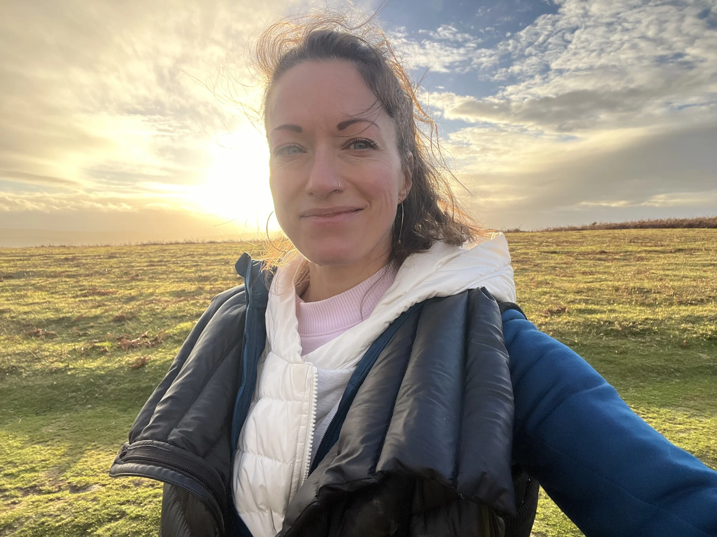 A woman with brown hair and blue eyes taking a selfie outdoors in a field during sunset. She has a slight smile, is wearing a black jacket with a hood, a white vest, and a pink shirt underneath.
