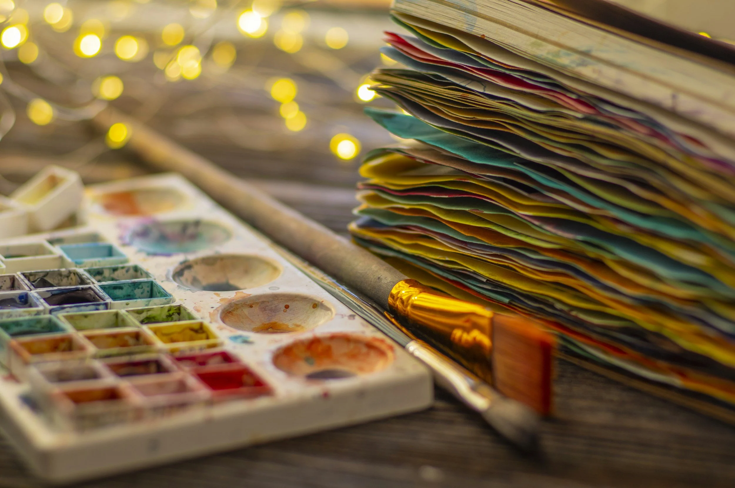 Colorful watercolor paint set, paintbrush, and stacked watercolor paper on a wooden surface with blurred yellow fairy lights in background.