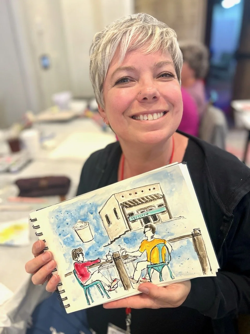 Woman with short gray hair smiling, holding a sketchbook with a colorful drawing of two people sitting at a table outside a coffee shop.