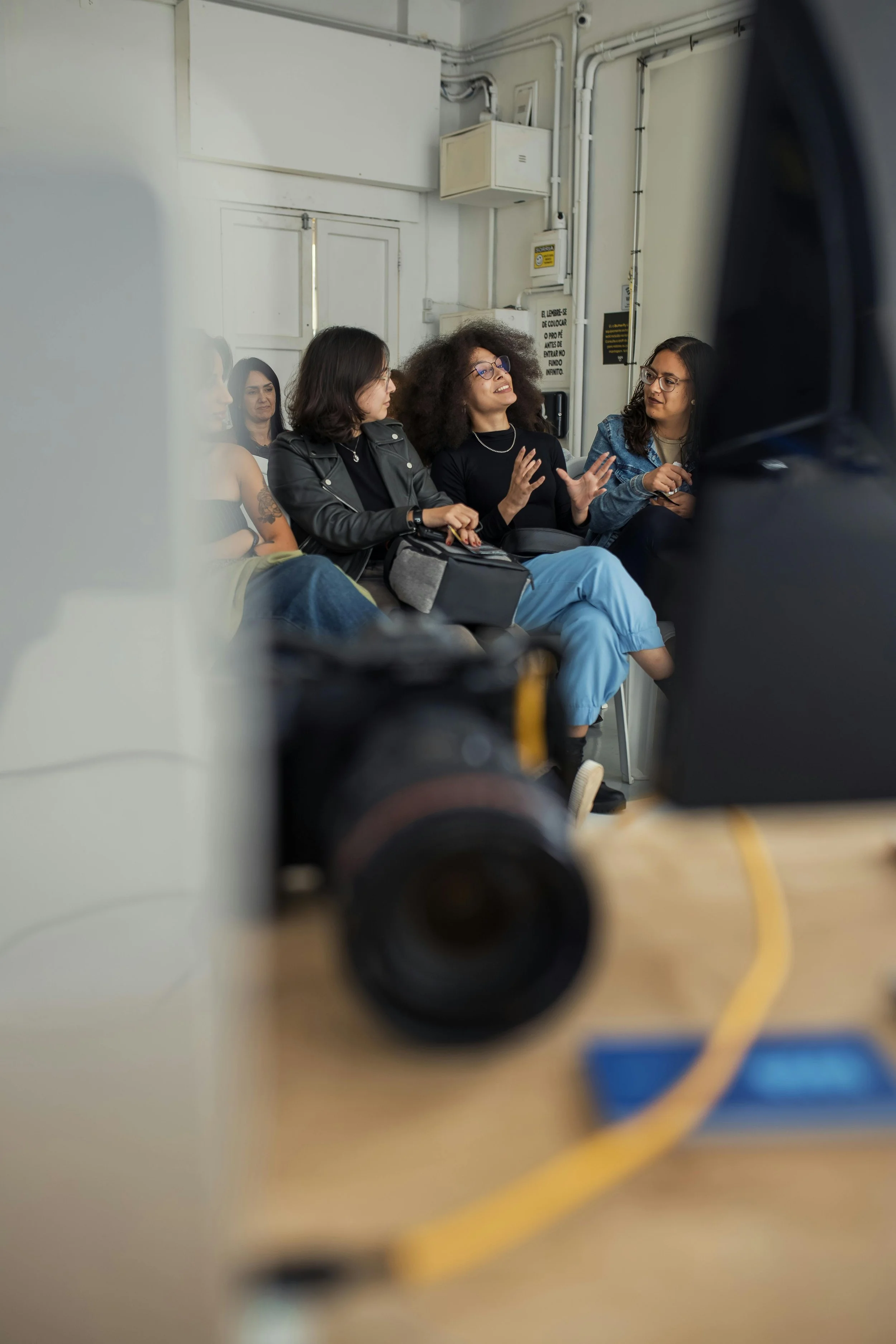 A group of women sitting and talking in a room with a white wall, one woman is gesturing with the hands as she speaks, and a camera is partially visible in the foreground.