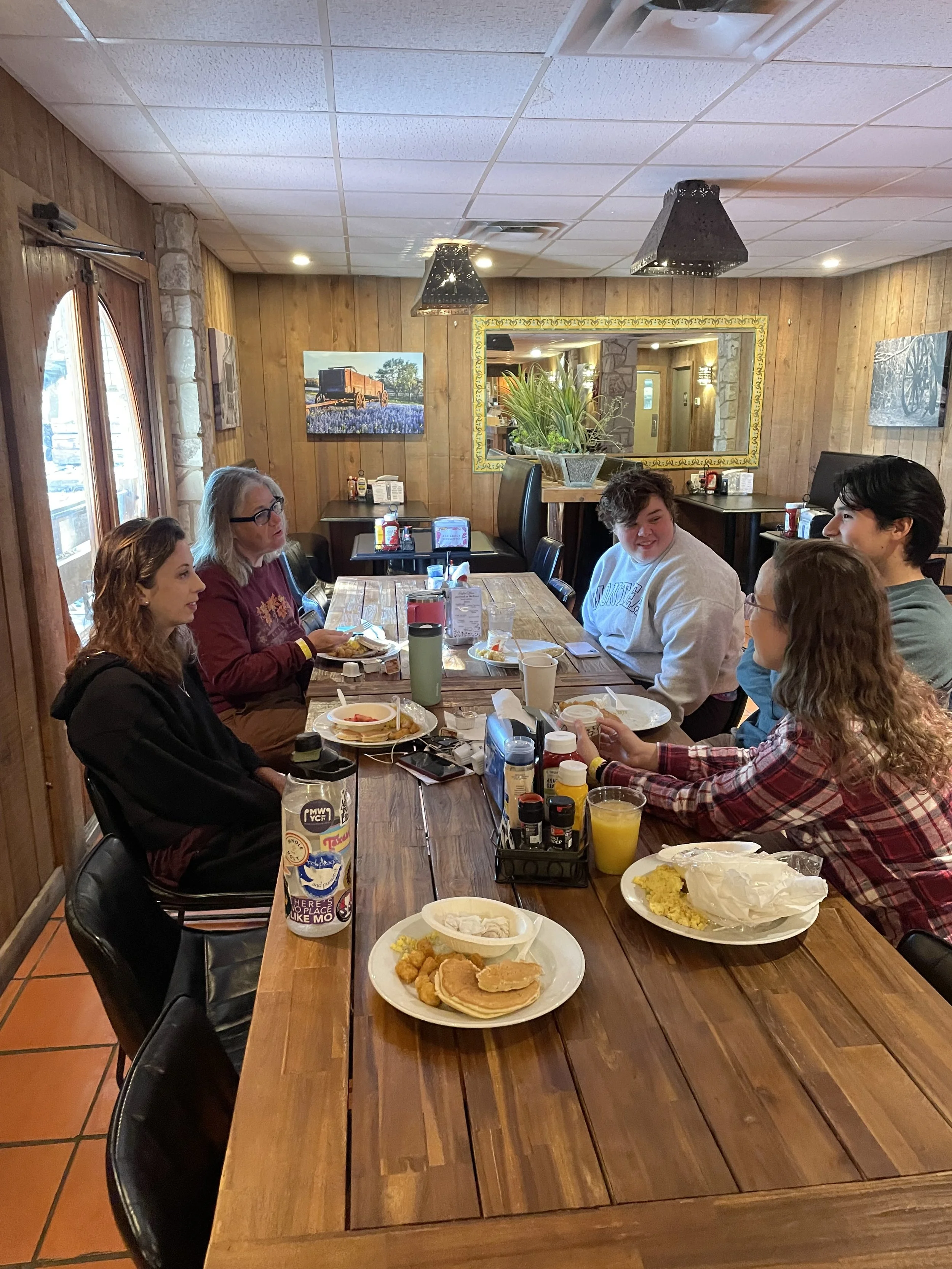 A group of five people sitting around a wooden table in a restaurant enjoying breakfast, with plates of pancakes, eggs, and breakfast foods, and drinks like juice and coffee.
