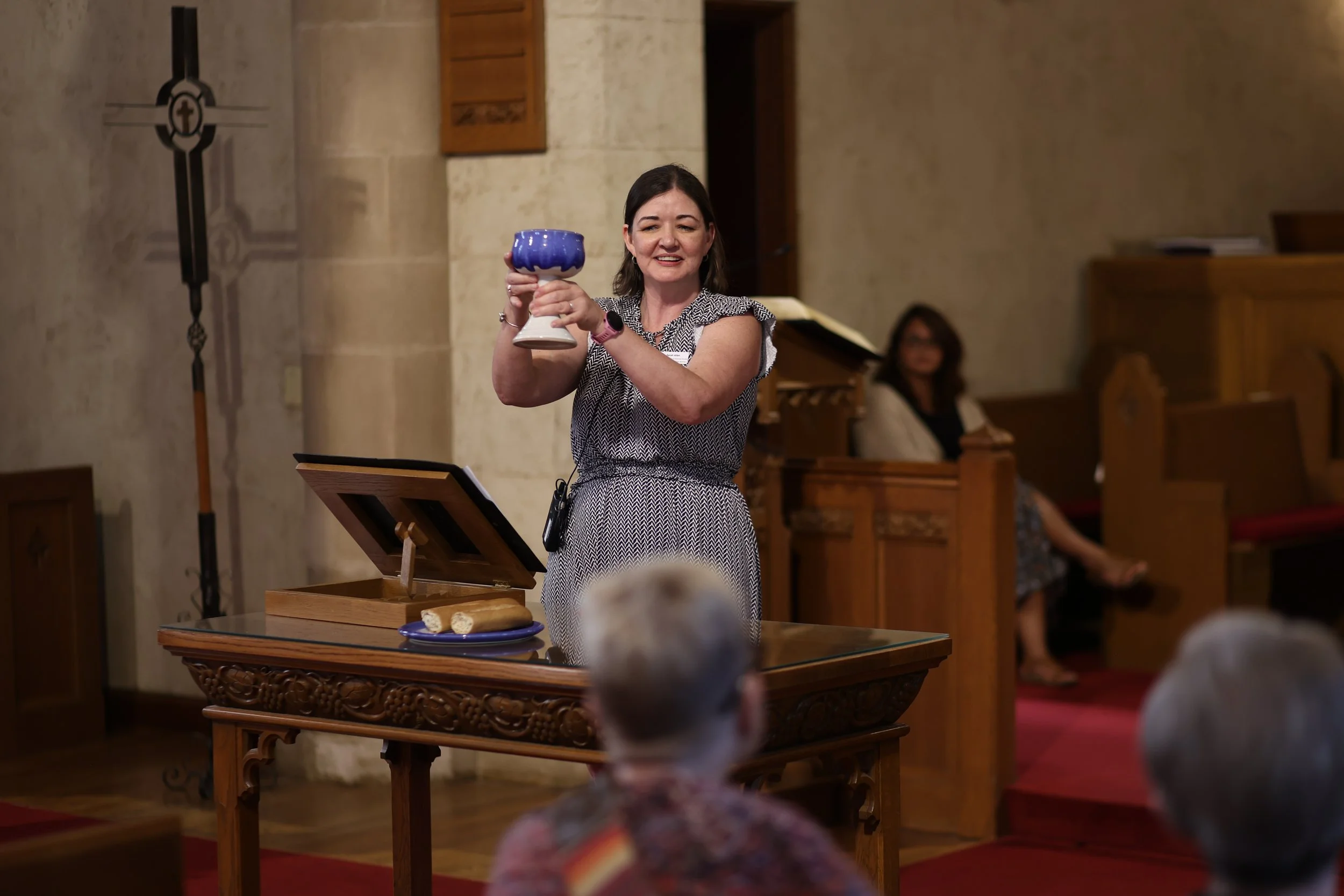 A woman standing behind a wooden table holding a blue chalice, inside a church with wooden pews and a stone wall, with two women seated in the background.