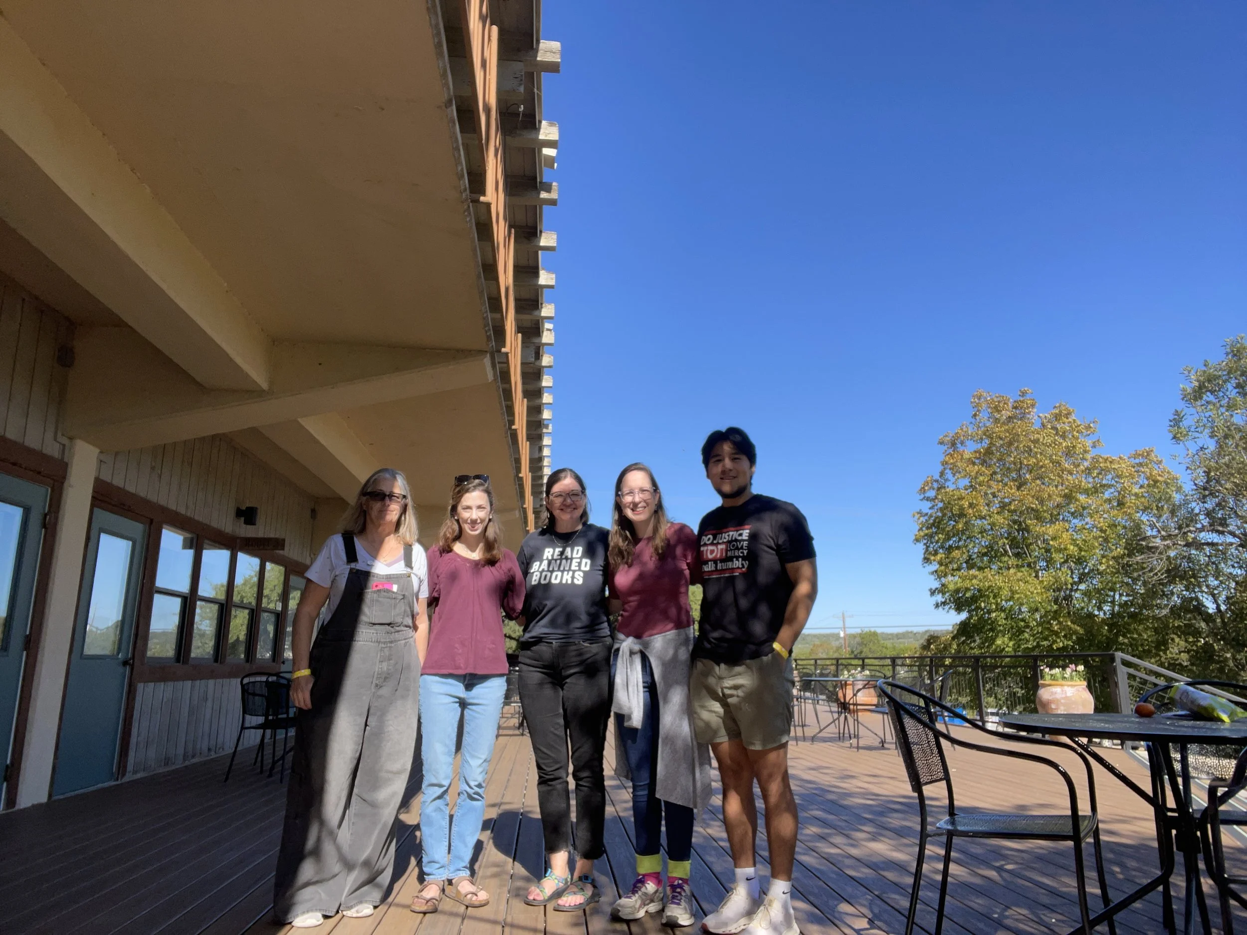 Five people standing on a wooden deck outside near a building with large windows, smiling at the camera with trees and a clear blue sky in the background.
