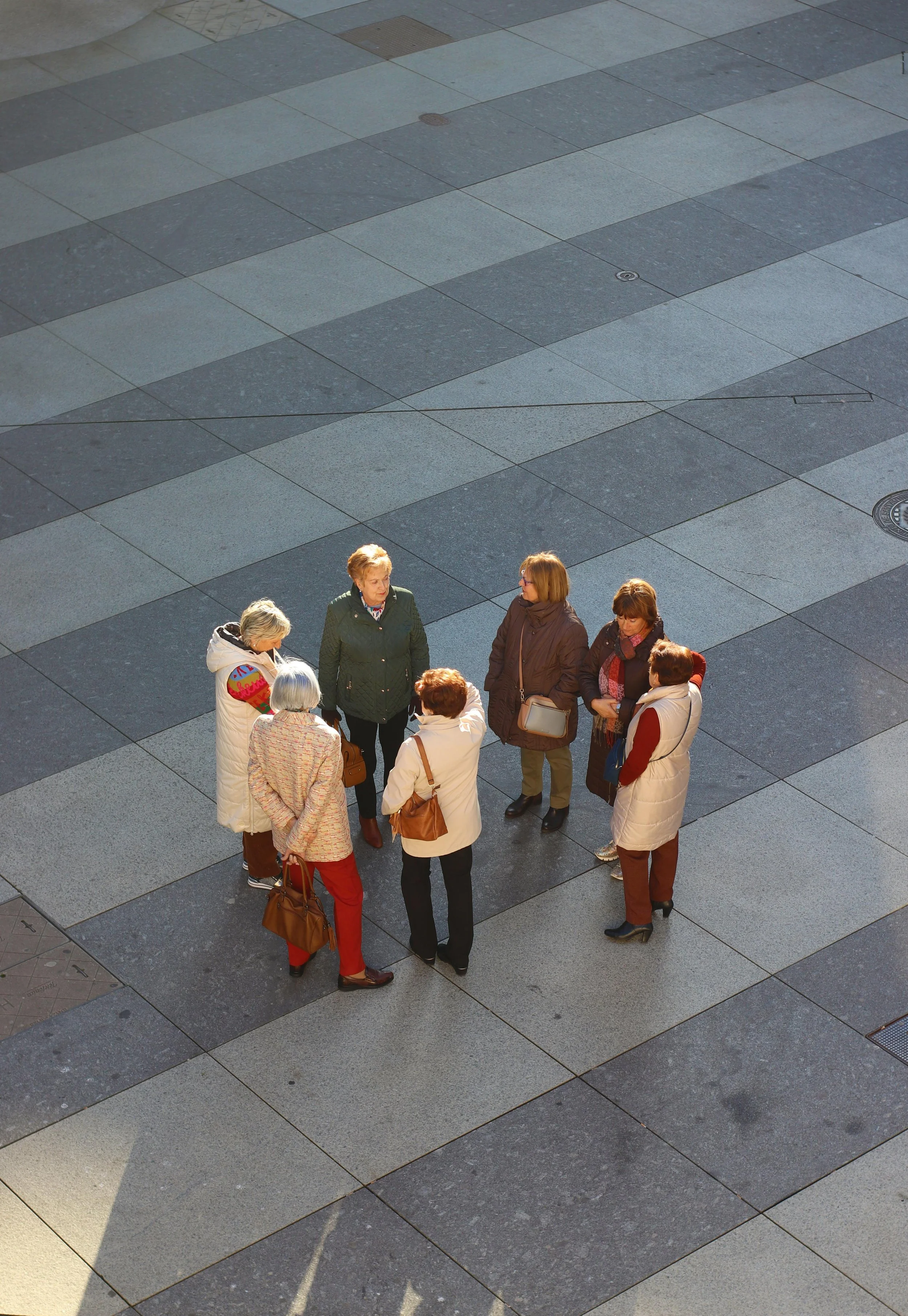 A group of seven women standing in a circle on urban pavement, engaged in conversation, with sunlight casting shadows.