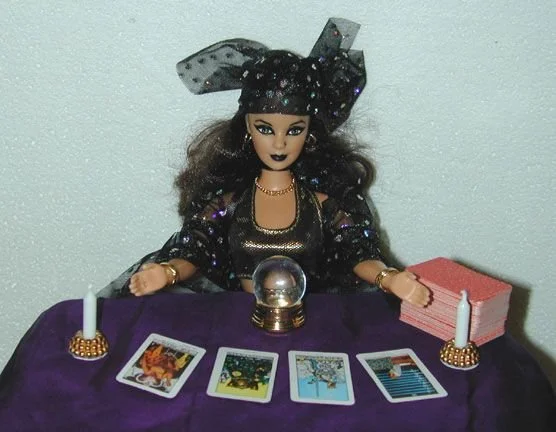 A Barbie doll with black hair sitting at a table and reading tarot cards.