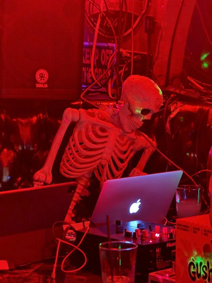 A skeleton propped as a DJ in front of a laptop and a drink.