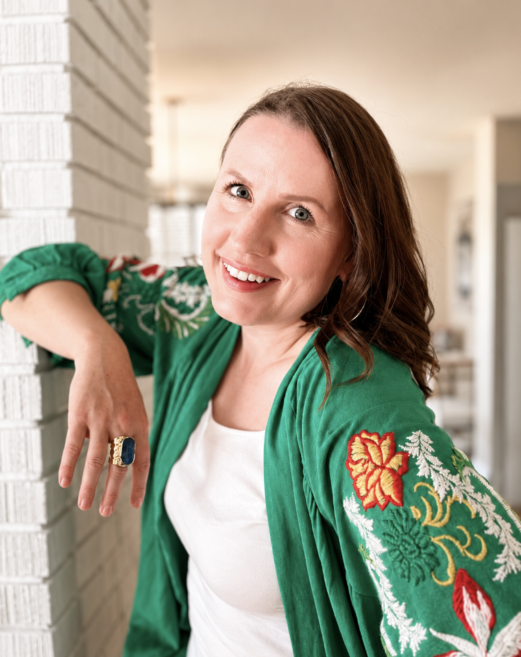 A woman with brown hair and blue eyes smiling, wearing a green embroidered jacket and a white shirt, leaning on a brick wall in an indoor setting.