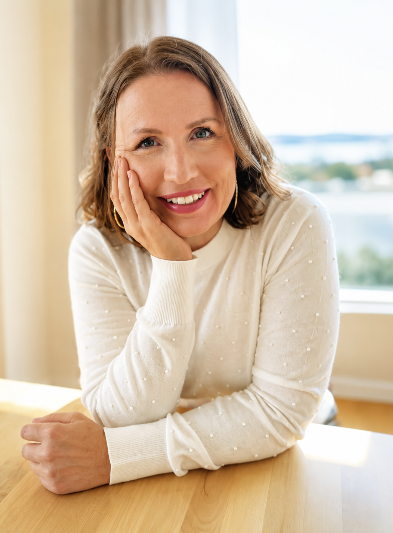 A woman with shoulder-length brown hair, wearing a white sweater with small pearl-like embellishments, smiling and resting her head on her right hand, sitting at a wooden table near a window with a blurred landscape view in the background.