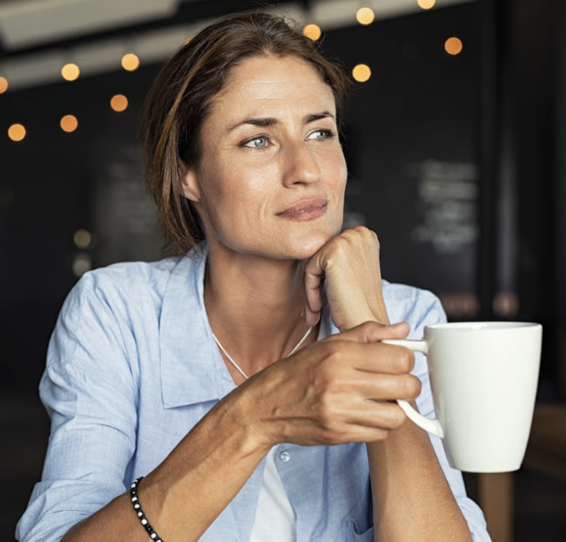 A middle-aged woman with short brown hair and blue eyes, wearing a light blue shirt, sitting in a cafe, holding a white mug in her right hand, appearing thoughtful as she gazes out of the frame with a soft, contemplative expression, with blurred string lights in the background.