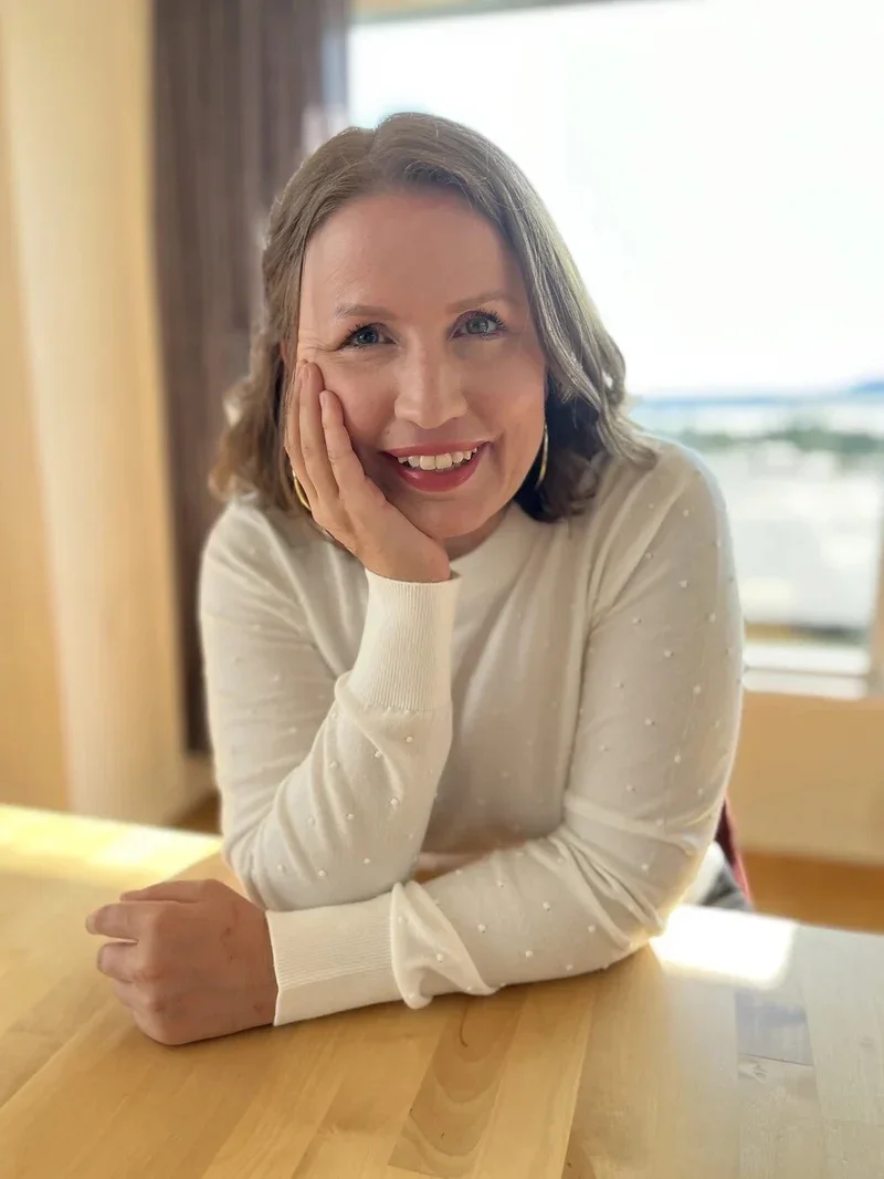 A woman with shoulder-length hair smiling and resting her face on her hand, sitting at a wooden table near a window with a view of the outdoors.