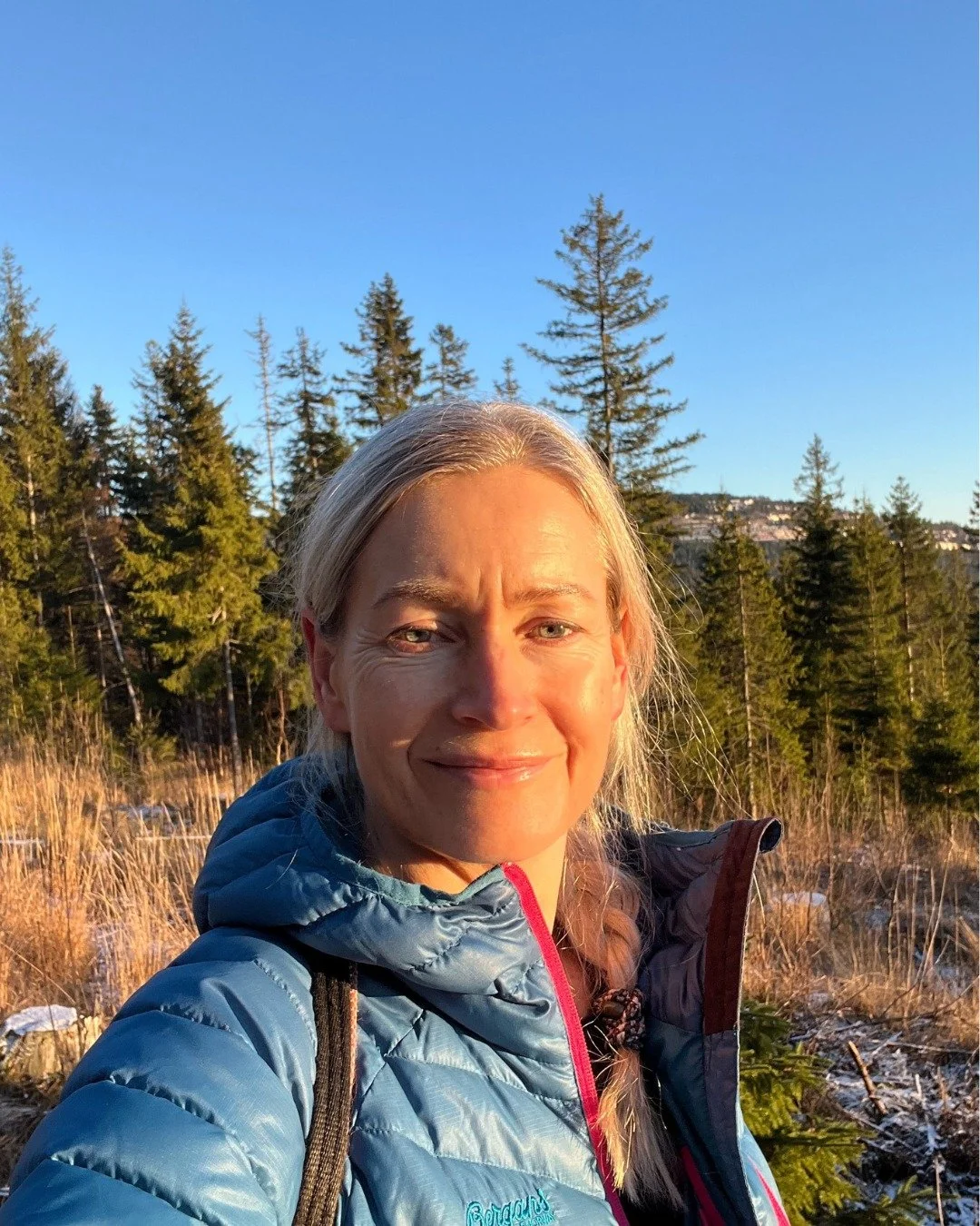 A woman smiling outdoors in a wooded area with tall pine trees and a clear blue sky, wearing a blue jacket and carrying a backpack.