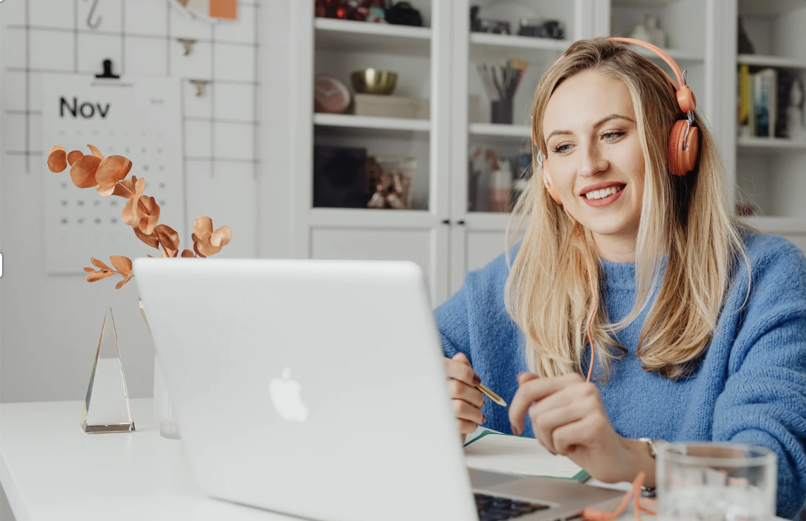 A woman with long blonde hair wearing a blue sweater and orange headphones smiling at her laptop while sitting at a white desk.