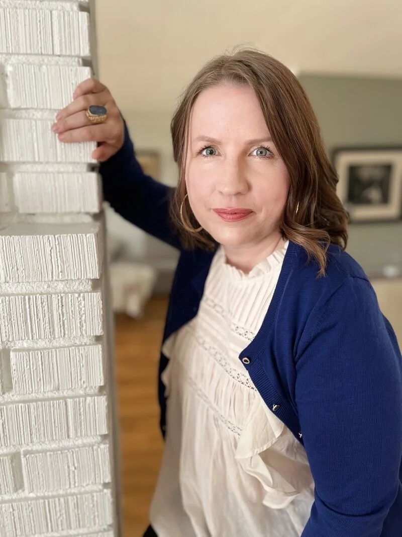 A woman with shoulder-length light brown hair, wearing a white blouse and a blue cardigan, leaning against a white brick wall inside a home, looking at the camera.