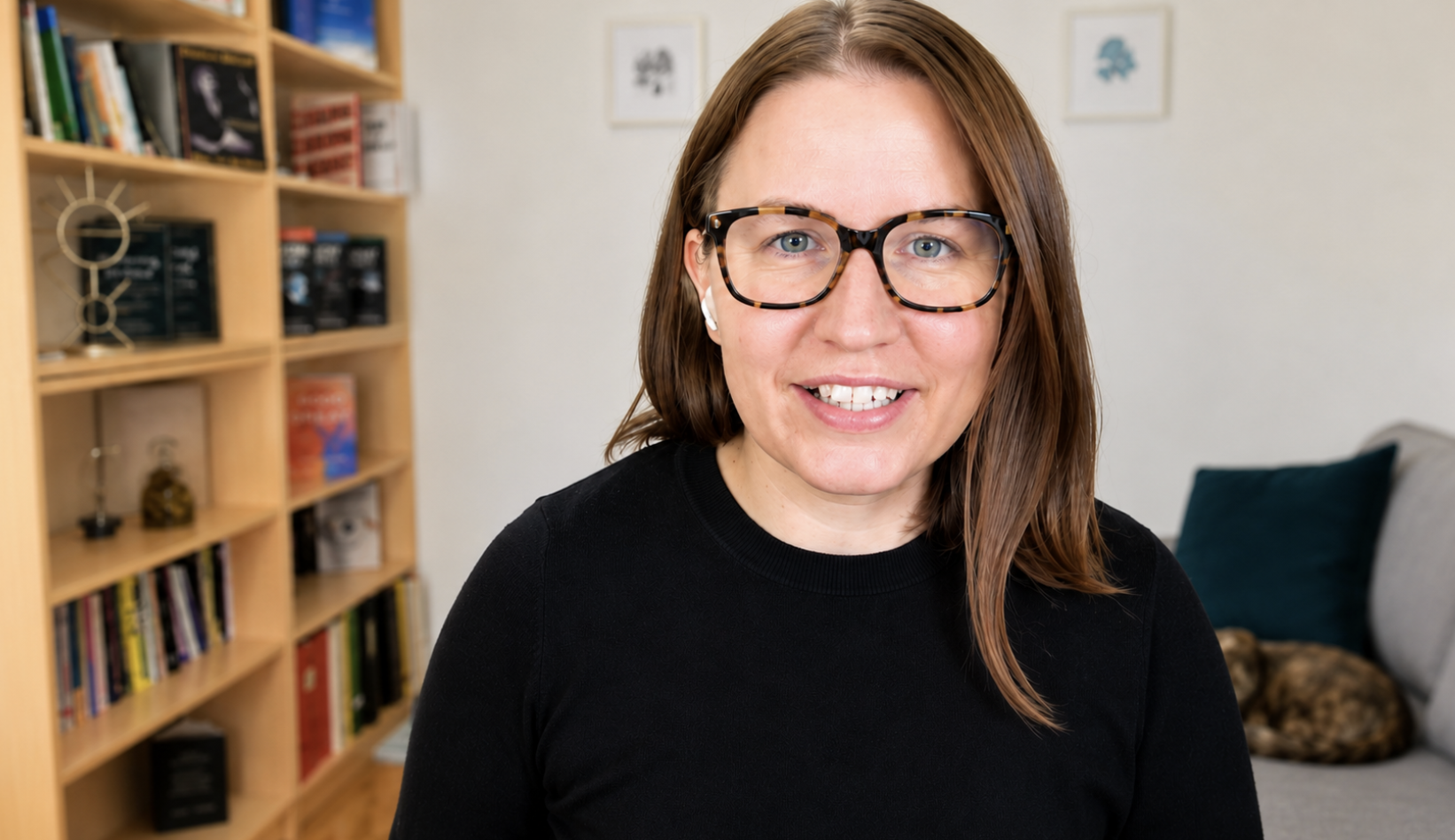 Woman with brown hair and glasses smiling in a room with a bookshelf and a dog sleeping on a couch in the background.