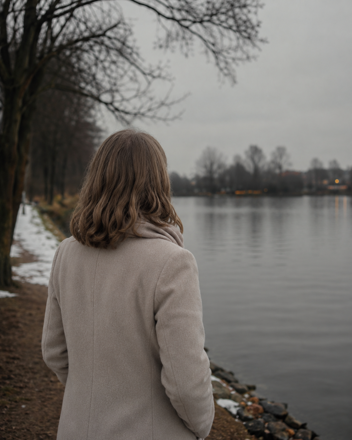 A woman with shoulder-length wavy brown hair wearing a light-colored coat walking beside a calm river on a cloudy, overcast day.