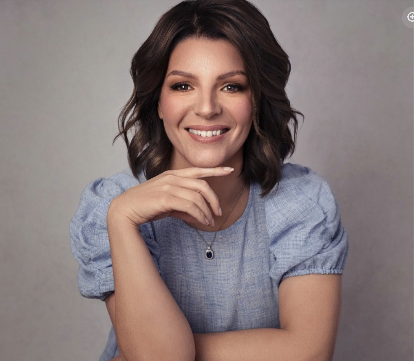 A woman with shoulder-length wavy brown hair smiling at the camera, wearing a light blue dress with puffed sleeves and a necklace with a dark stone pendant, her hand resting under her chin, against a plain gray background.