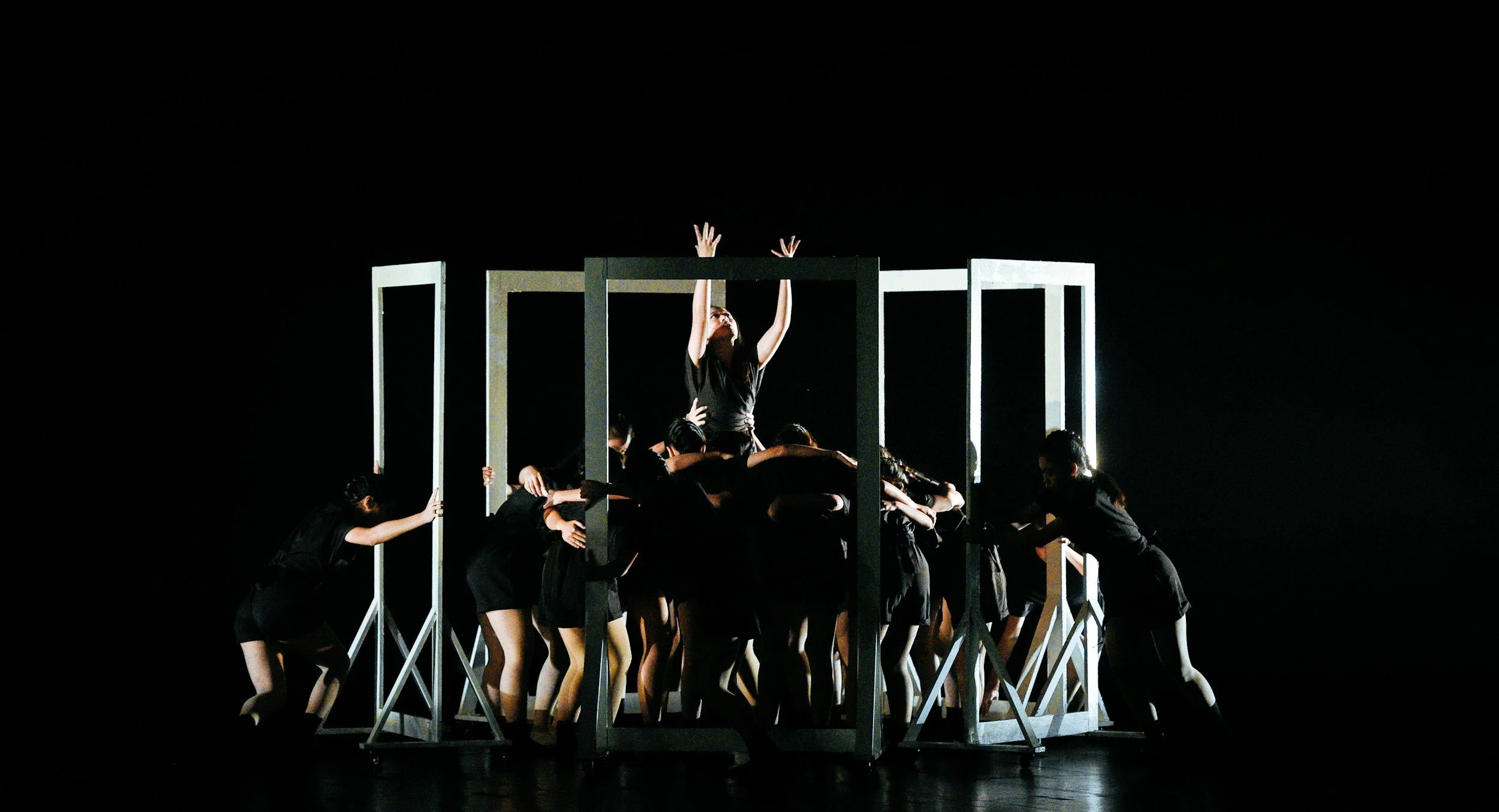 A group of performers in black costumes are surrounding a woman who is standing on a platform with her arms raised and head tilted back. The scene is dark with dramatic lighting highlighting the woman and the platform.