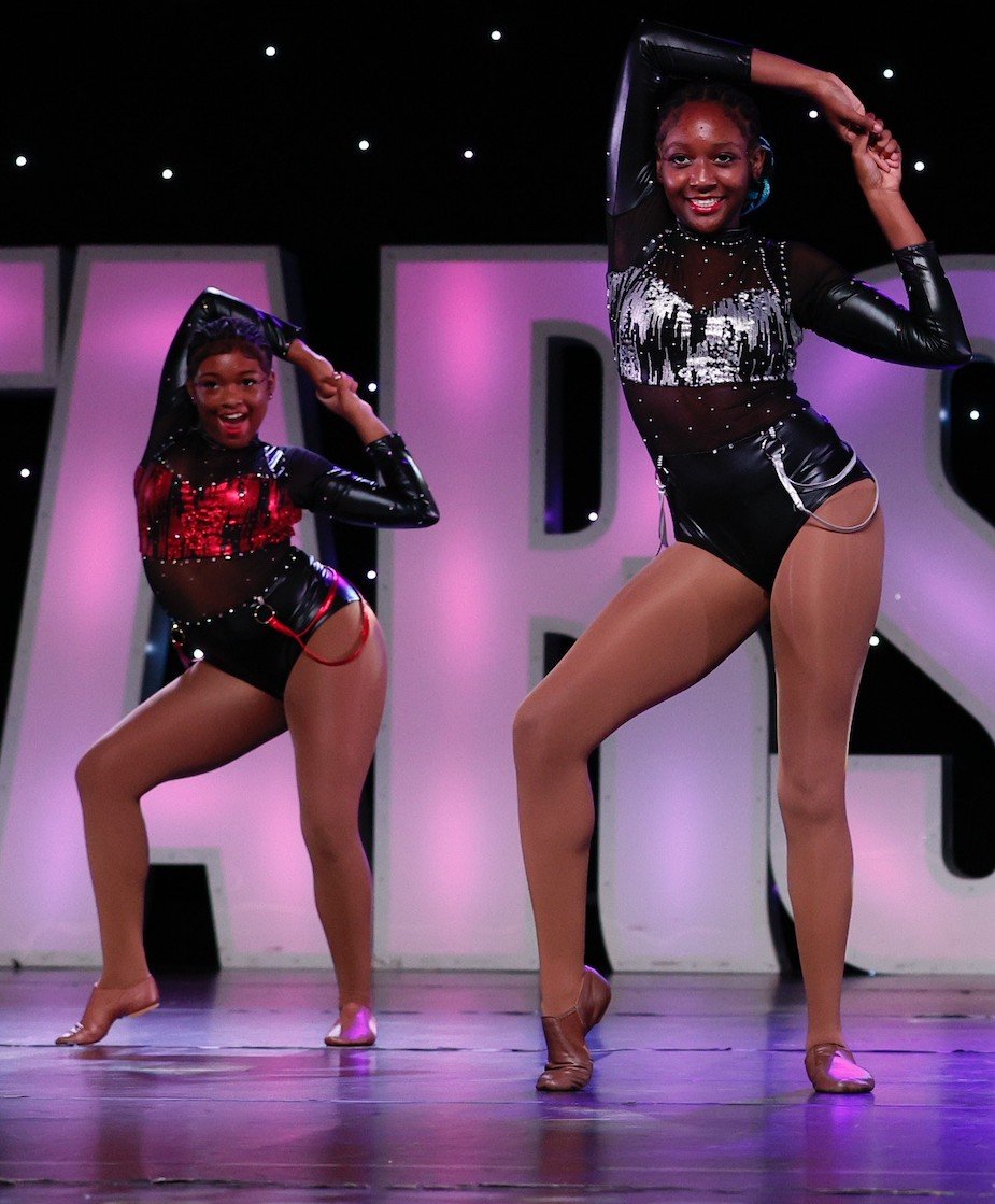 Two young girls performing a dance routine on stage, wearing black and red sparkly costumes with sheer tops and shorts, with stage lights and a large letter in the background.