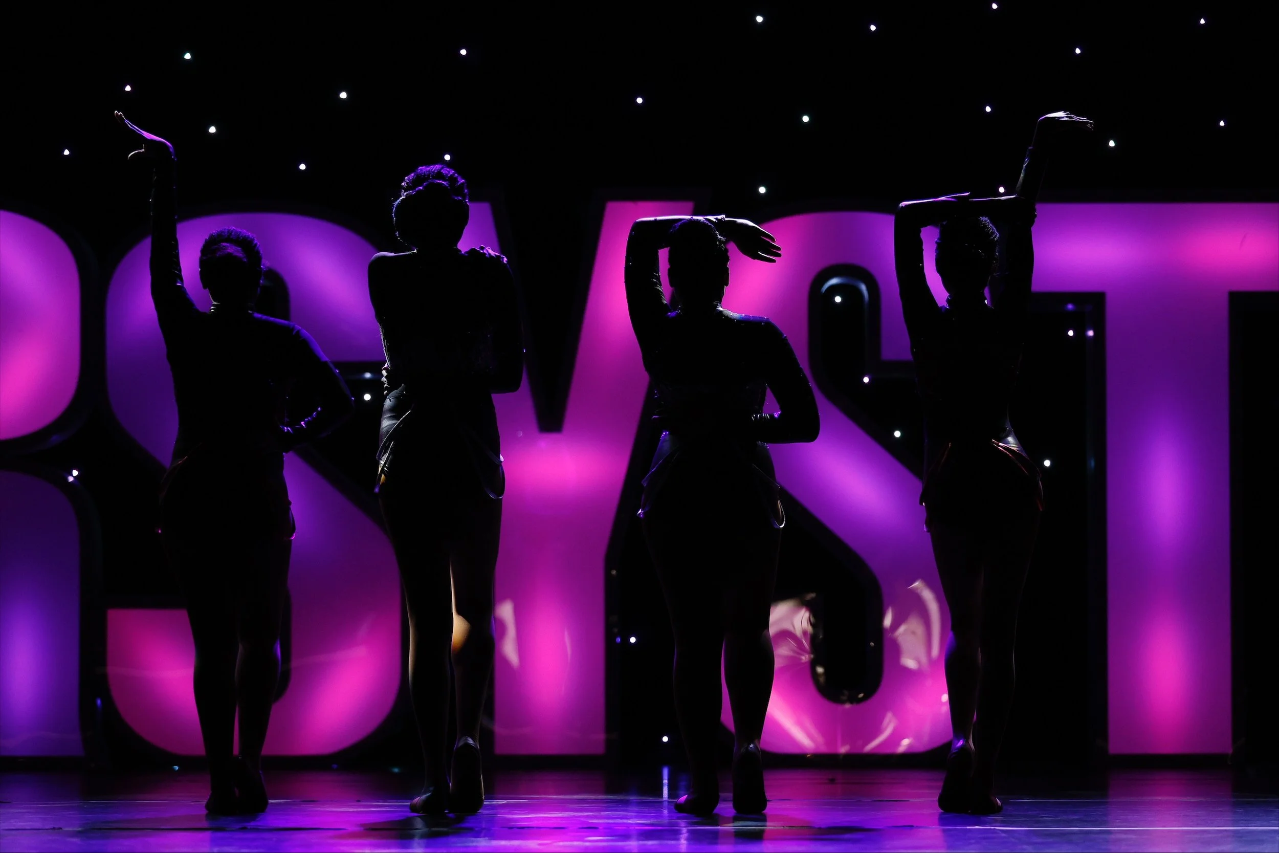 Silhouettes of four female dancers on stage with purple lighting, in front of large illuminated letters.
