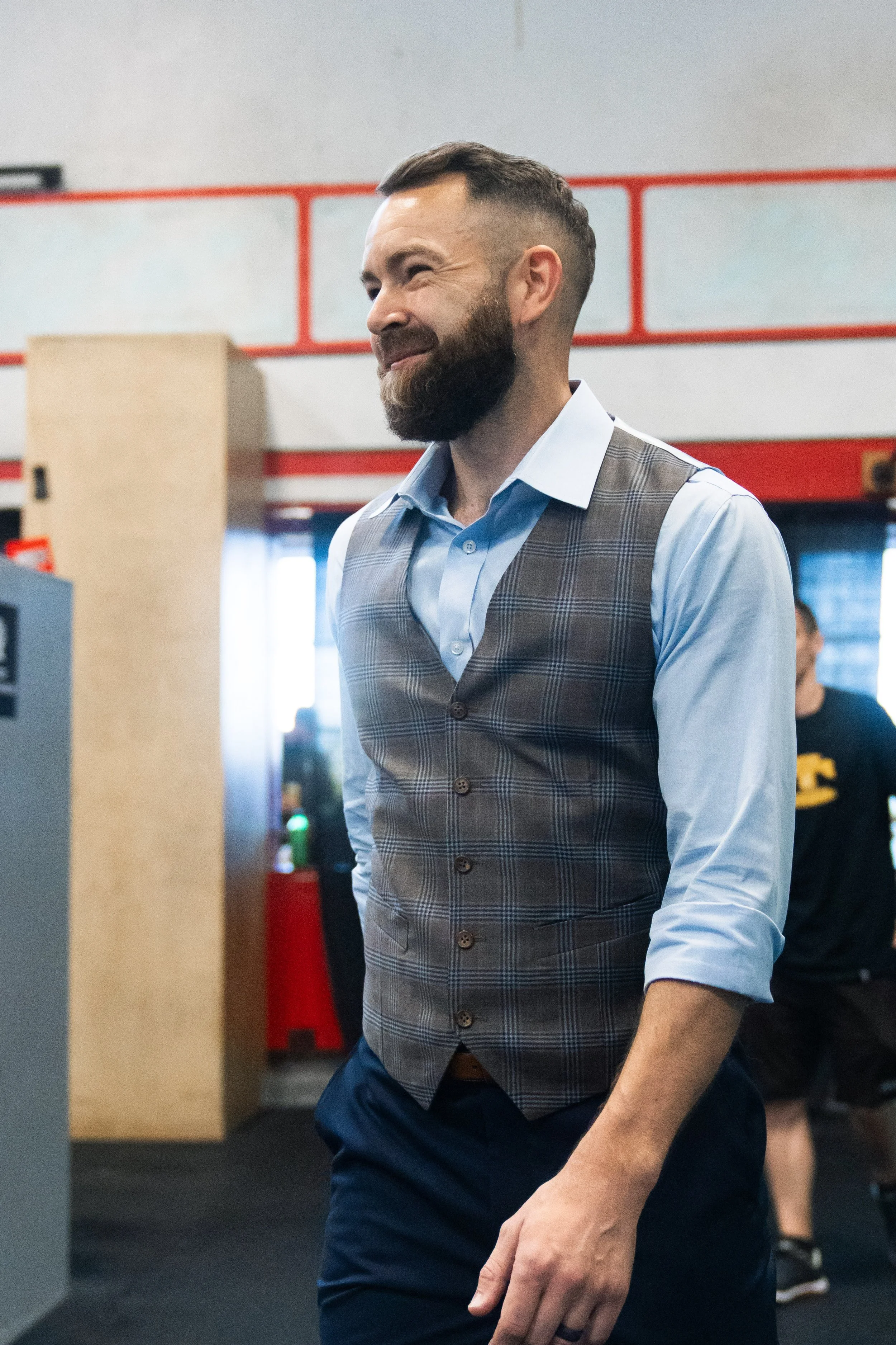 A man with a beard and a short haircut, wearing a light blue shirt and a brown plaid vest, smiling while standing indoors.