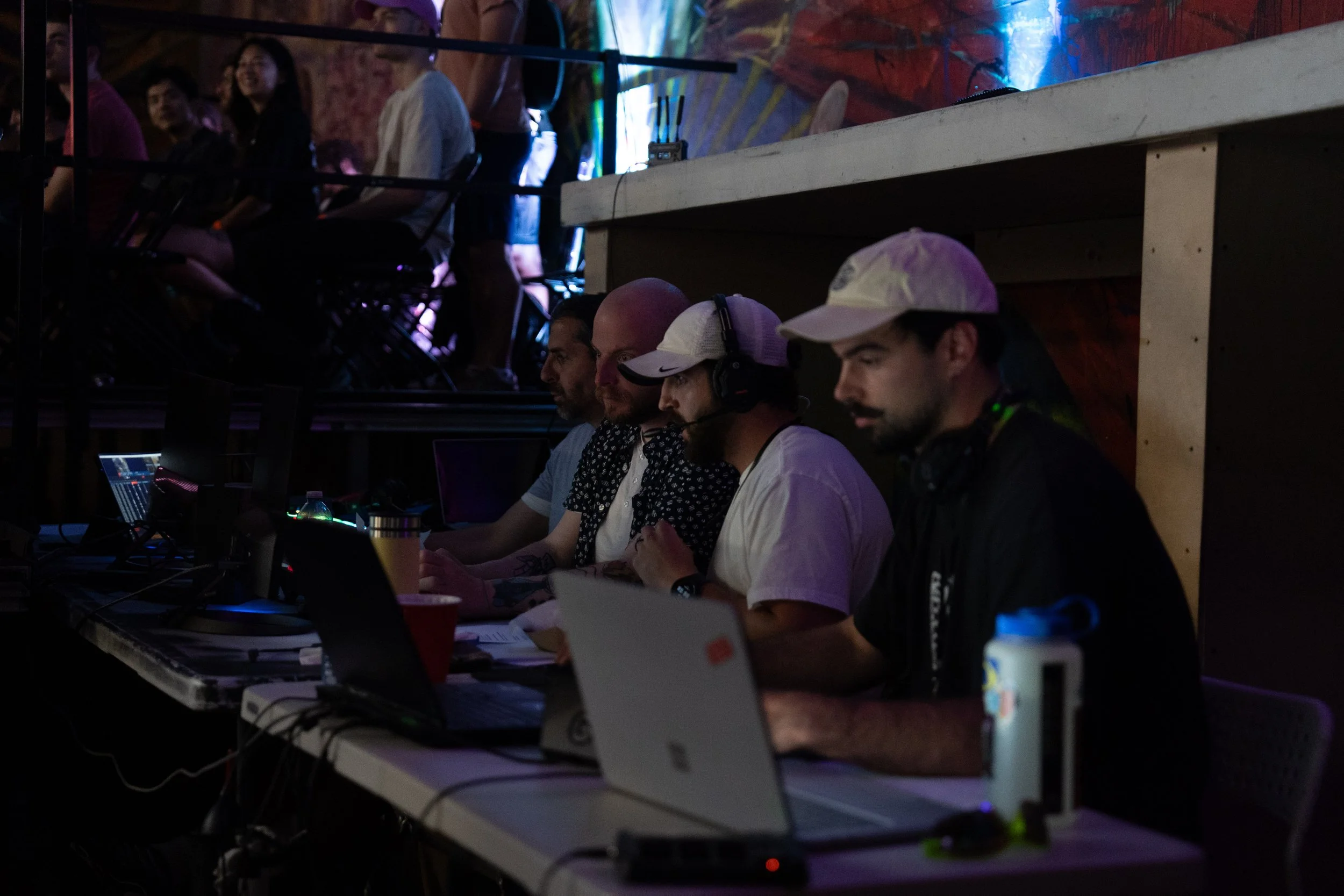 Group of men working on laptops at a table with equipment in a dimly lit, lively environment.