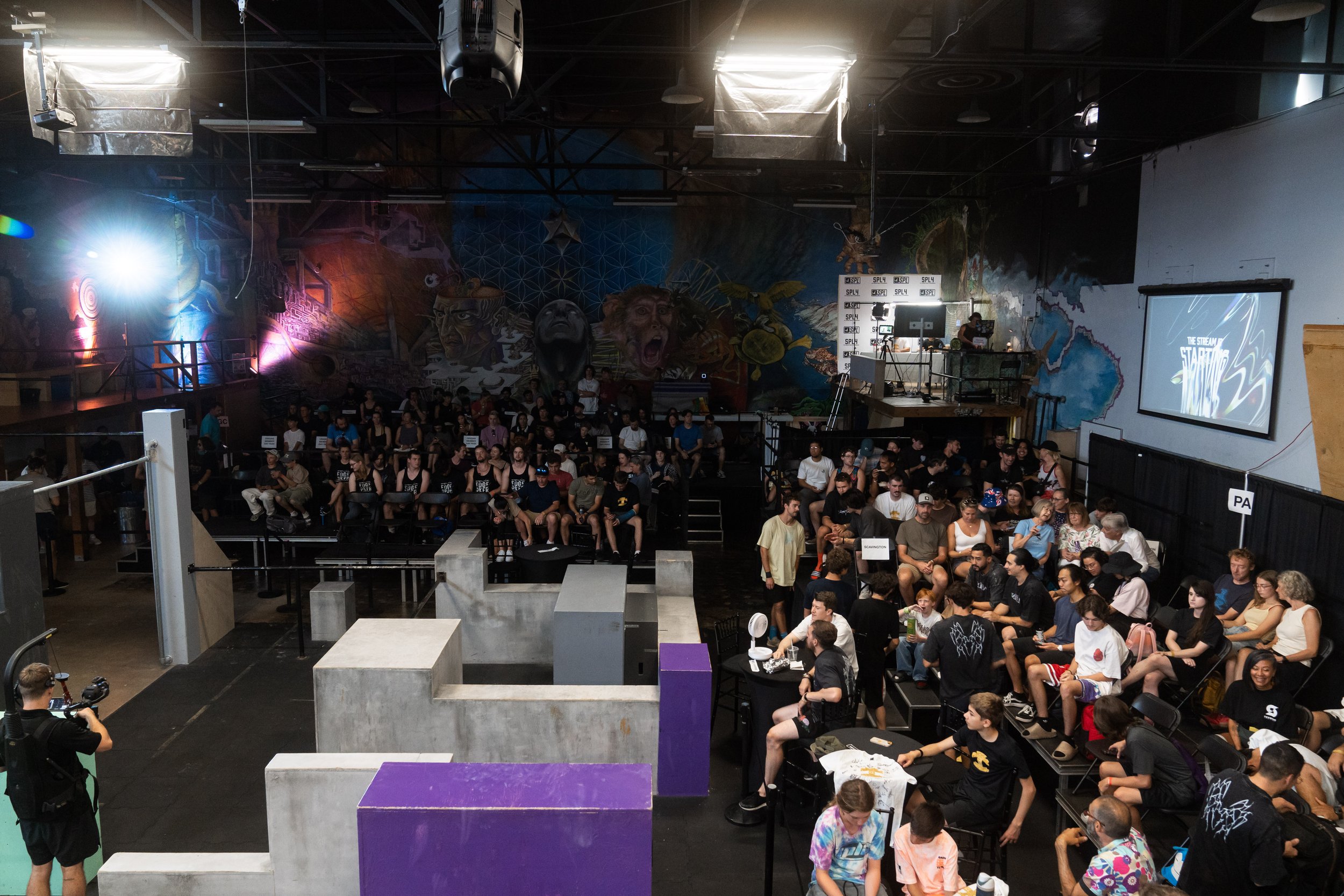 Audience gathered in a dark venue for a gaming or streaming event, with a large screen displaying 'STARTING SOON' and a colorful mural on the back wall.