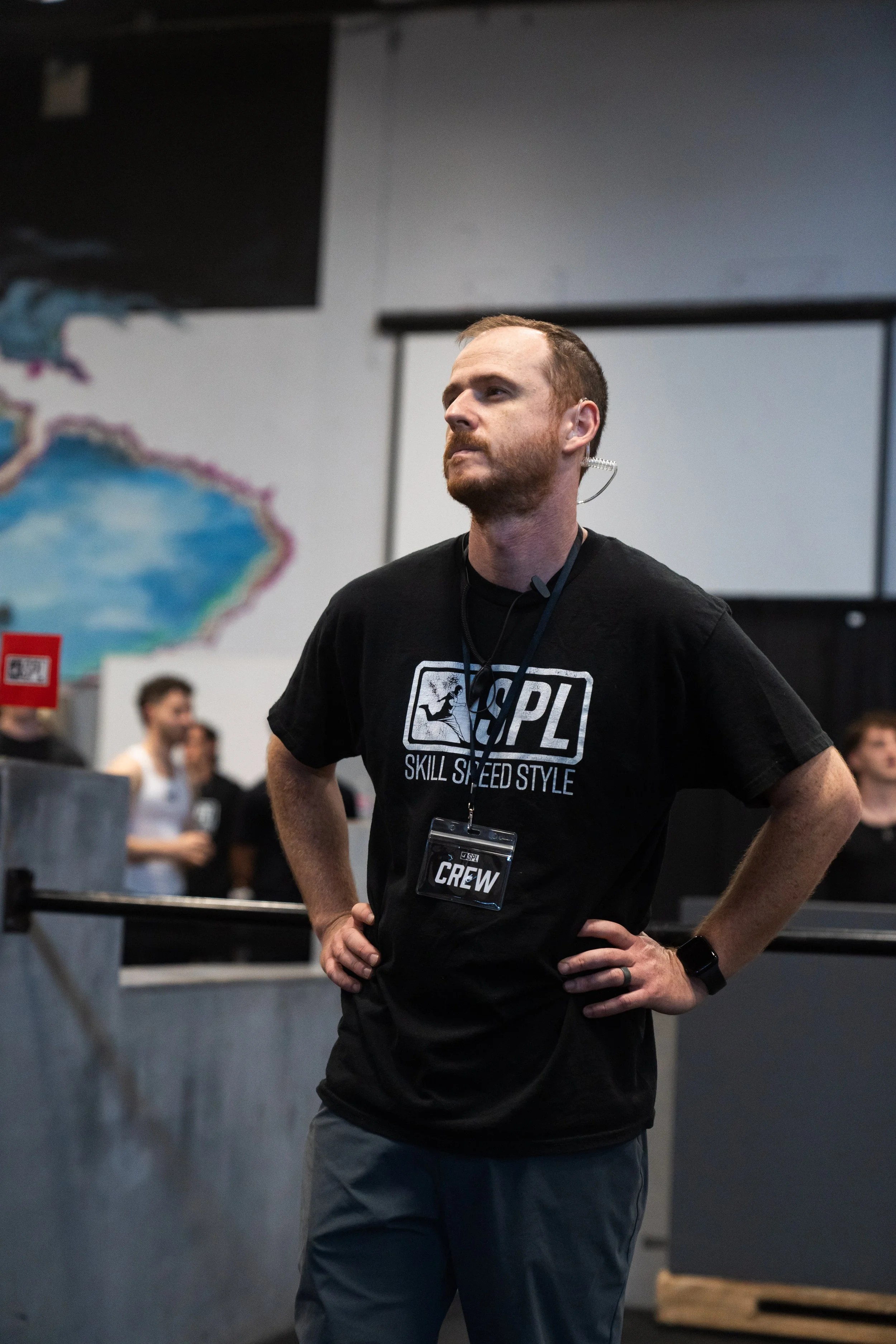 A man with a beard standing with hands on hips, wearing a black PL T-shirt with 'Skill Speed Style' logo, in a gym or fitness center, with other people and equipment in the background.