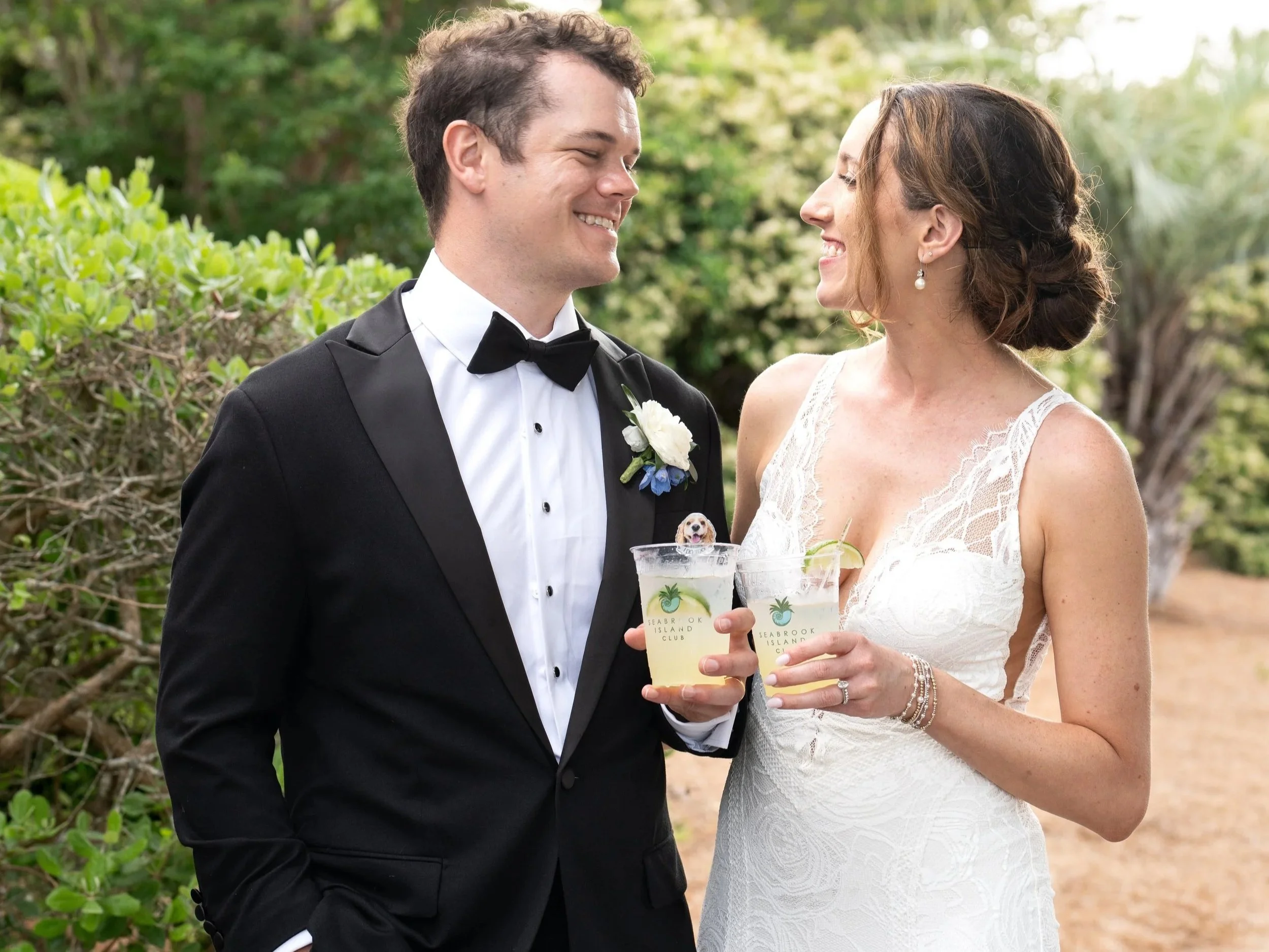 A bride and groom wearing wedding attire are smiling at each other while holding cocktails from Seabrook Island Club. The groom is wearing a black tuxedo with a bow tie, and the bride is in a white lace wedding dress with pearls.