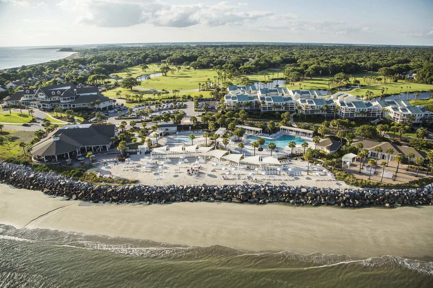 Aerial view of the beachfront Seabrook Island Club with a pool area, beach chairs, and umbrellas, situated along a sandy shore with a rock barrier and ocean, surrounded by lush greenery and a golf course in the background.