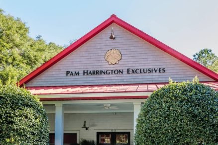 Front view of the Pam Harrington Exclusives office with a red gabled roof, white siding, and sign reading "Pam Harrington Exclusives" with a shell logo above.